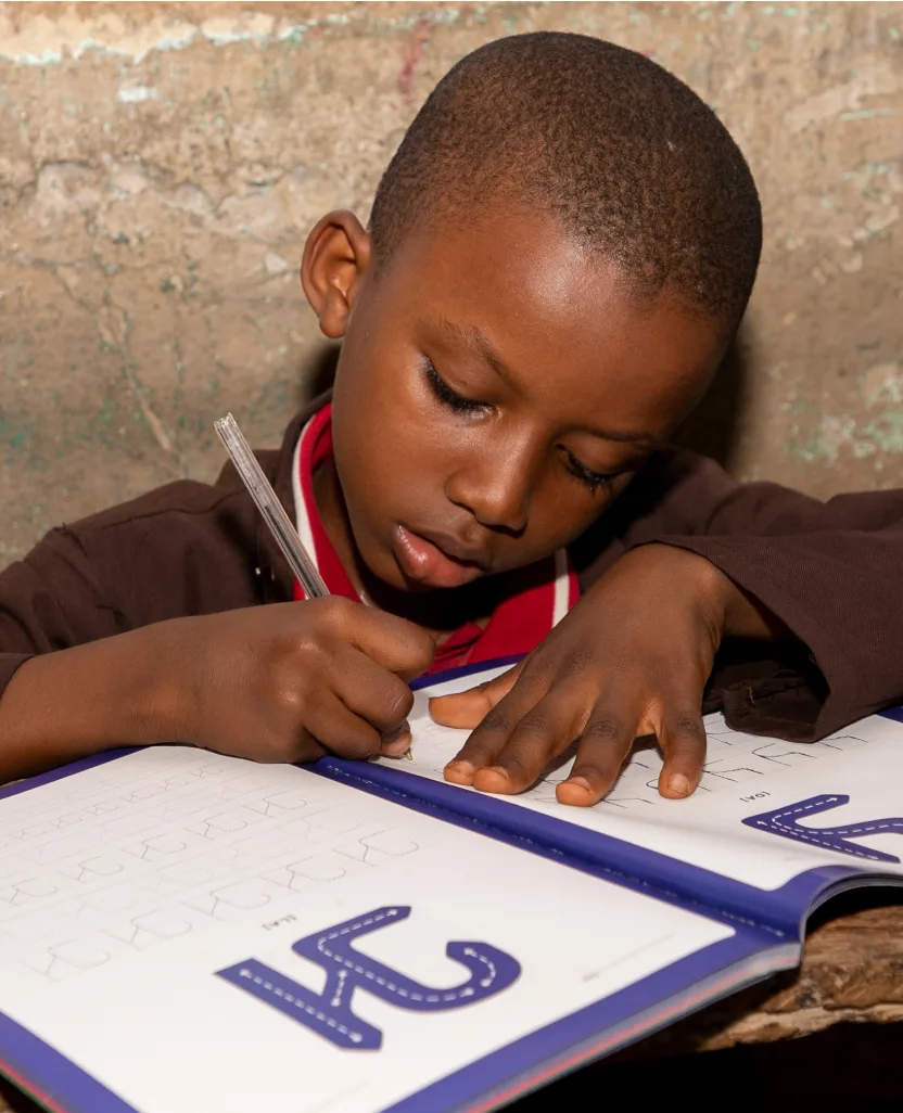 A young boy in a brown shirt with a red collar holds down a book with big blue ADLaM letters on the pages as he writes in it using a pen.