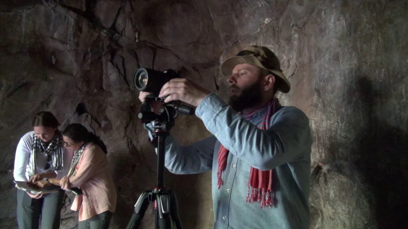 in a cave a man looks into a camera and two people look at a book