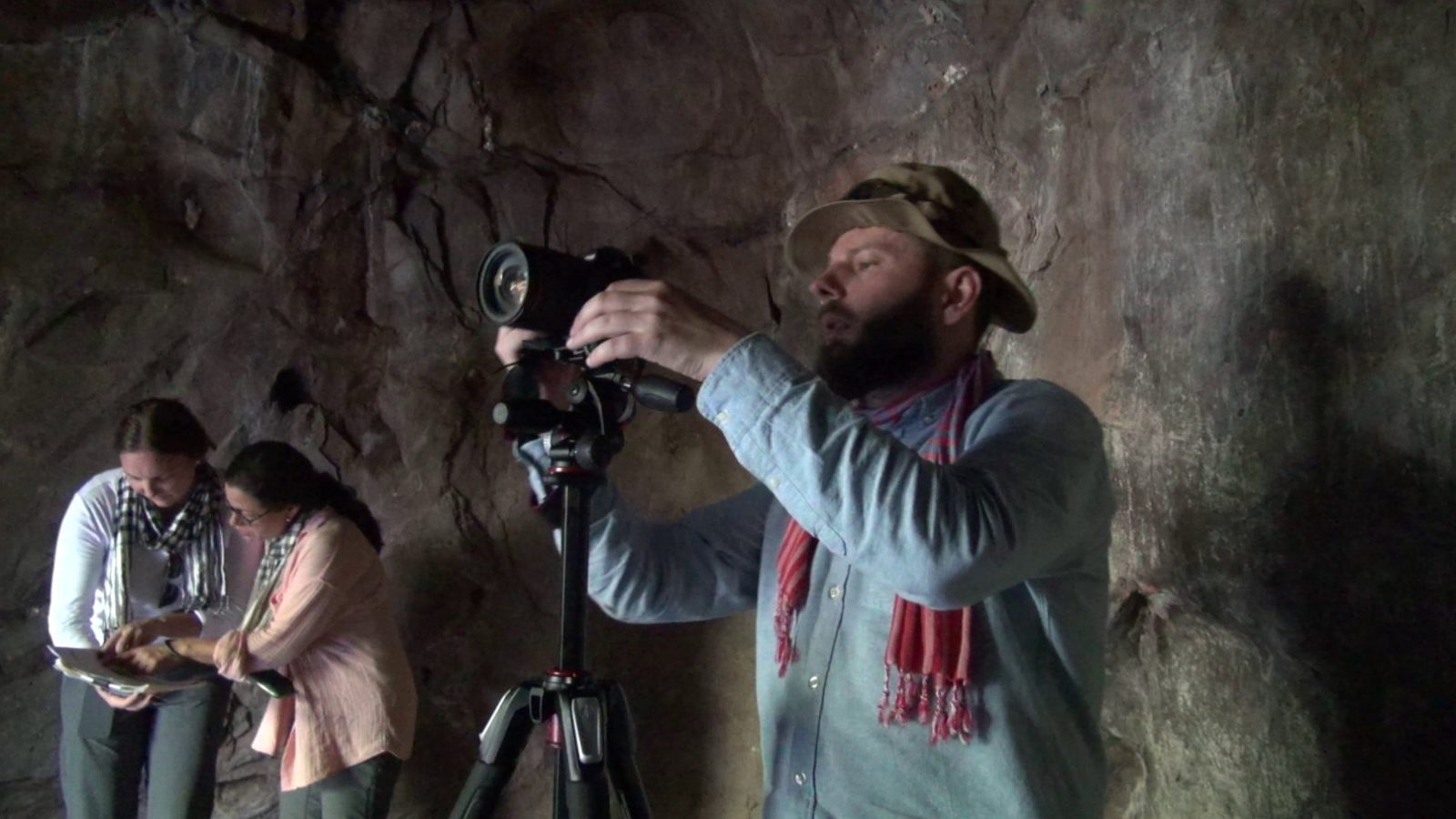 in a cave a man looks into a camera and two people look at a book