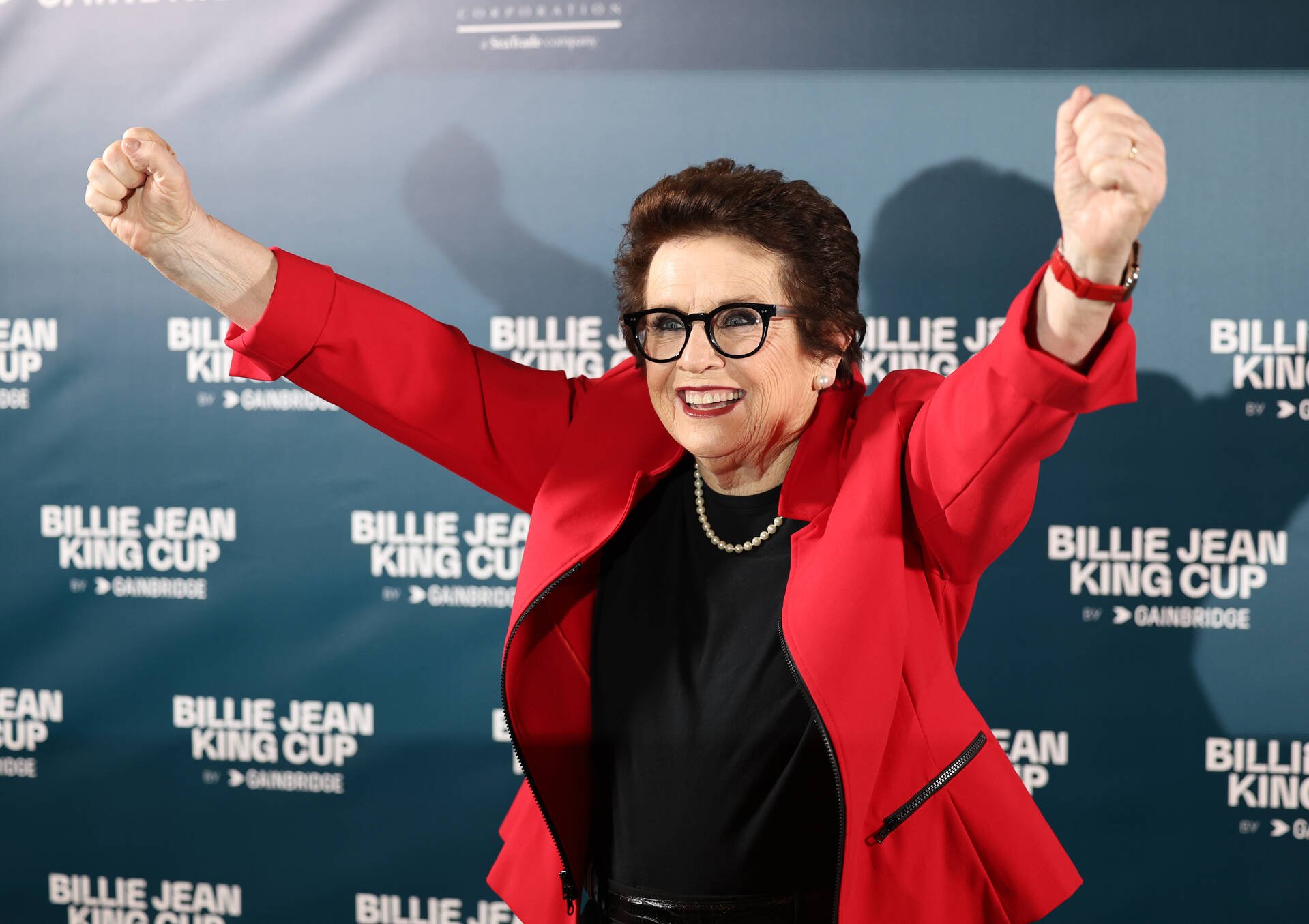 Person in a red jacket with raised arms, smiling in front of a backdrop with "Billie Jean King Cup" text.