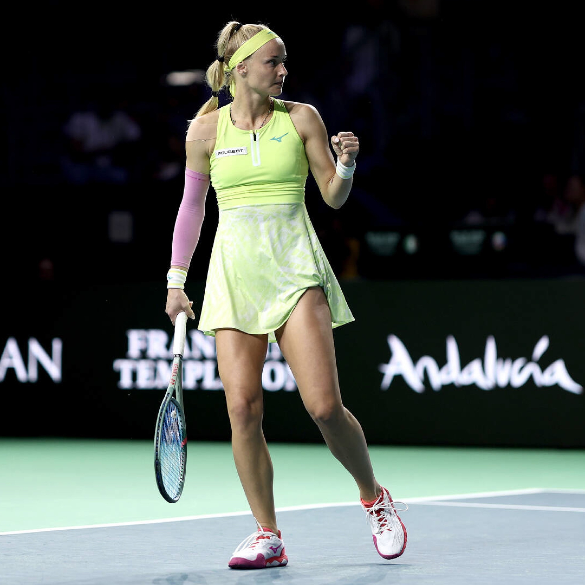 Tennis player in a light green outfit pumps fist on court, holding a racket, with a black background featuring advertisements.