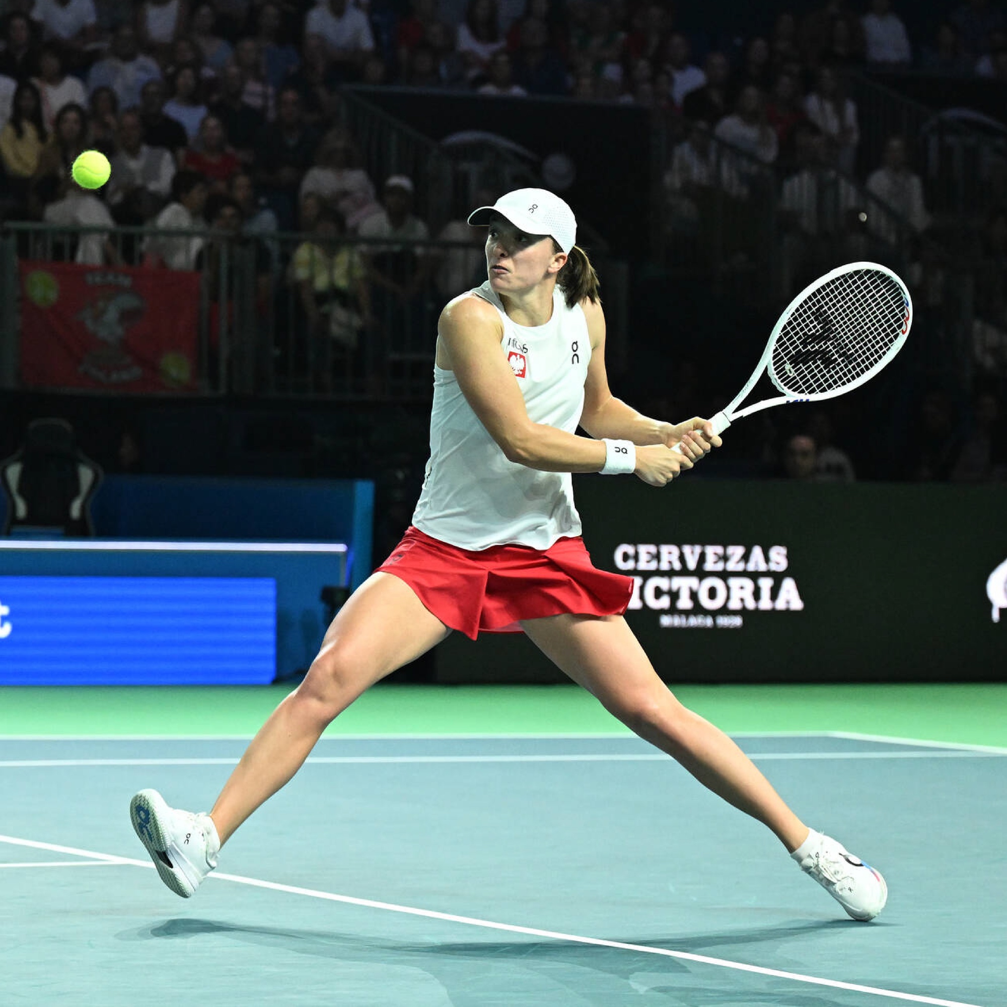 Tennis player in white top and red skirt hits a two-handed backhand shot during a match on a hard court.