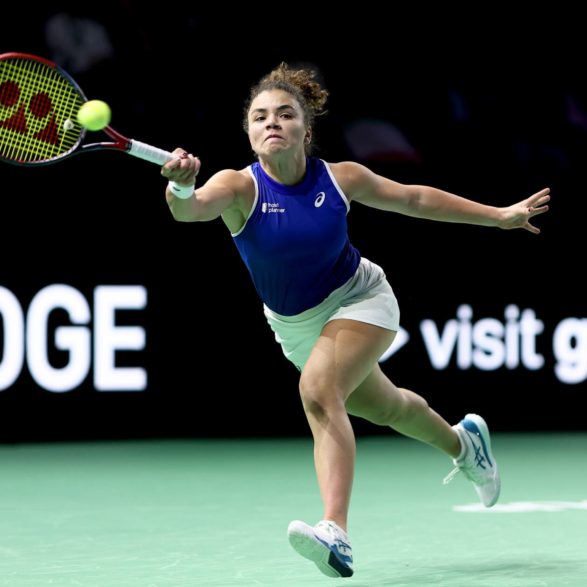 Tennis player in a blue shirt and white skirt lunges to hit a ball with a racket on a green court.