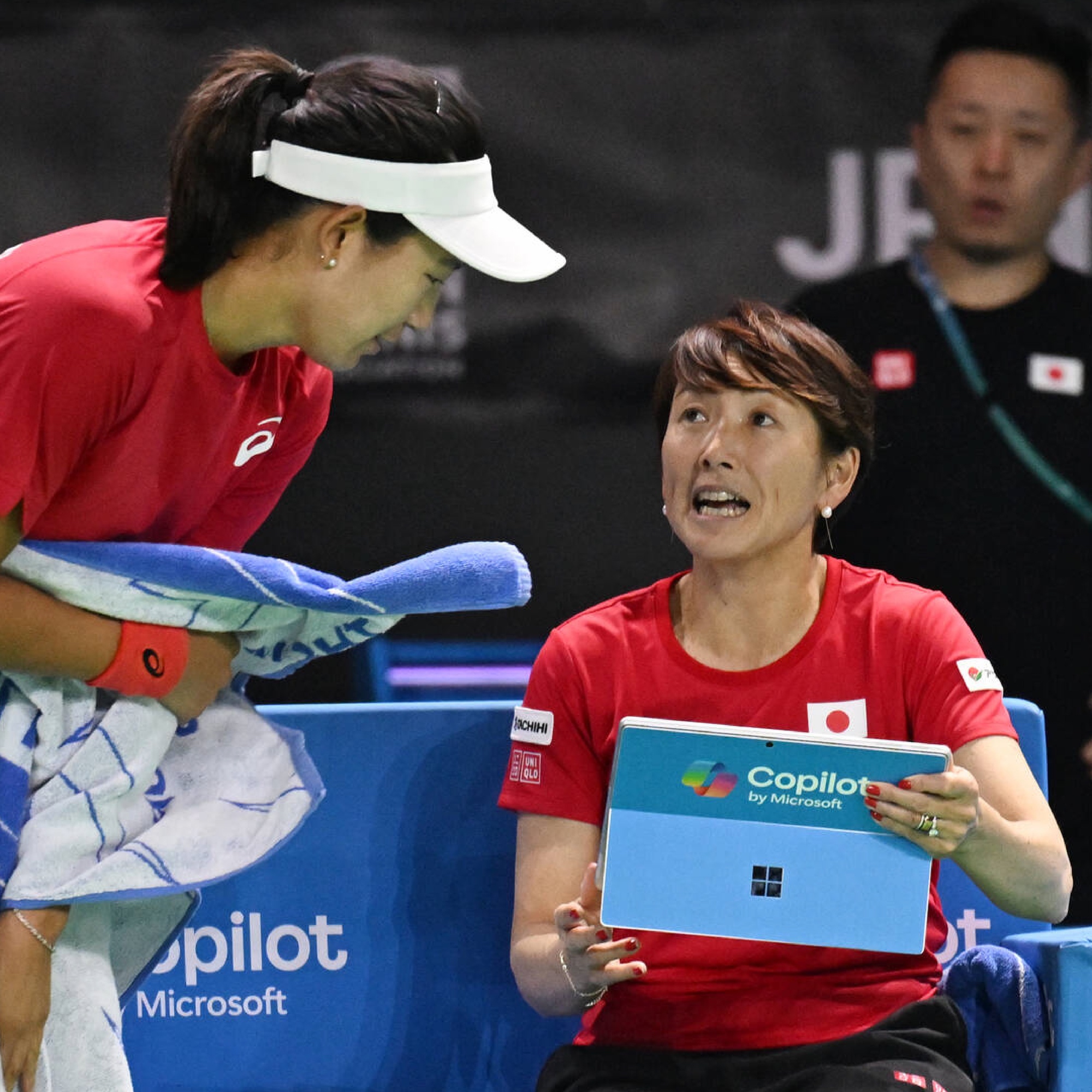 Two women in red shirts, one standing with a towel, the other sitting with a tablet. They seem to be having a conversation during a sports event.