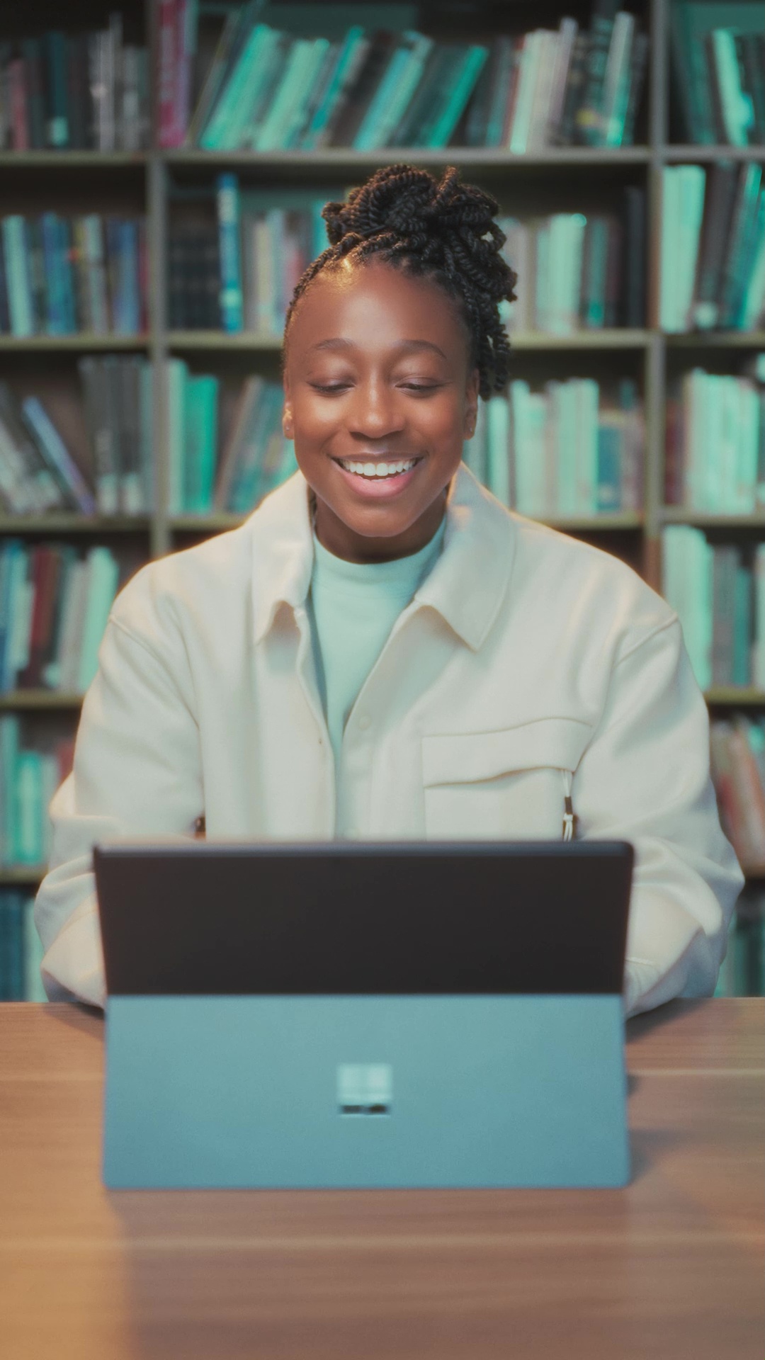 A person sits at a table smiling while using a laptop, with bookshelves filled with books in the background.