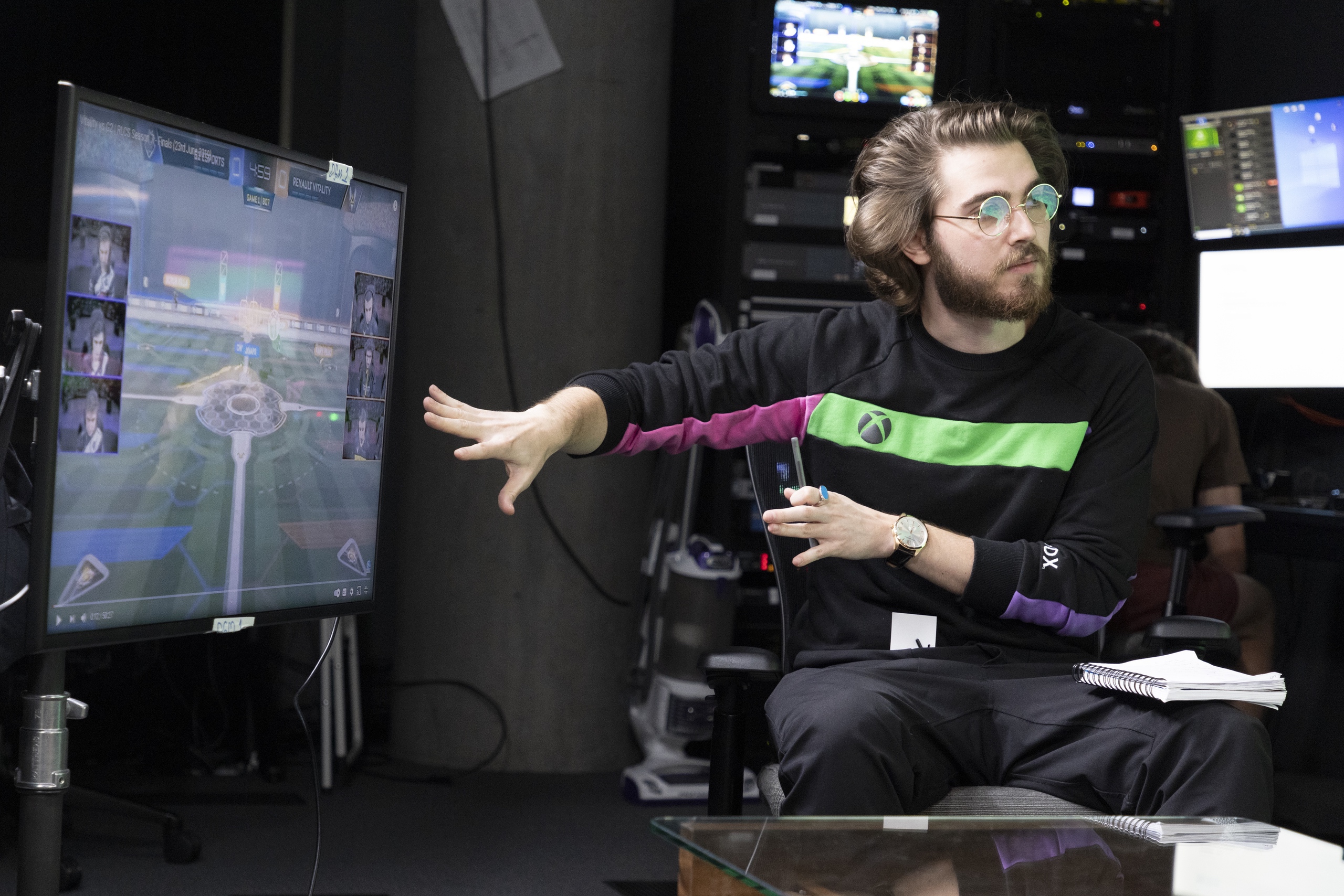 A person wearing glasses is sitting in front of a screen with his hands up and notepad on his lap. There are many screens behind him and to the side of him. His shirt is black and has an XBOX logo in green.
