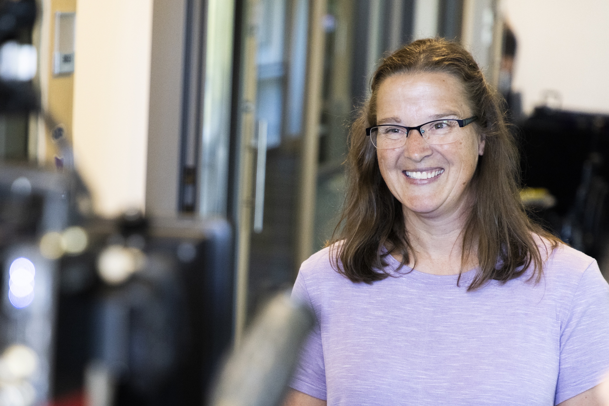 A woman is standing and smiling on the right side of the image. She is wearing glasses and a purple shirt. In the bottom left foreground of the image, a blurry camera is visible.