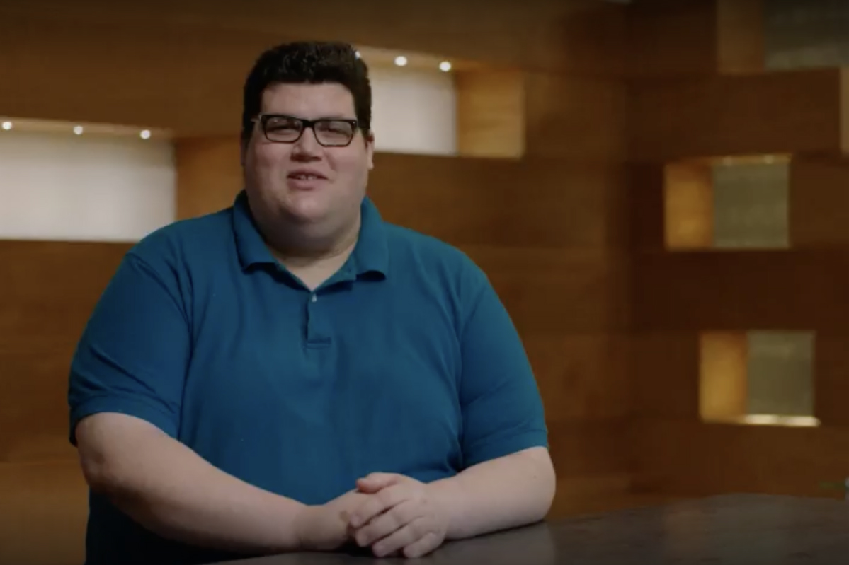 A man in a blue shirt and glasses leaning on a table in front of a wood background.