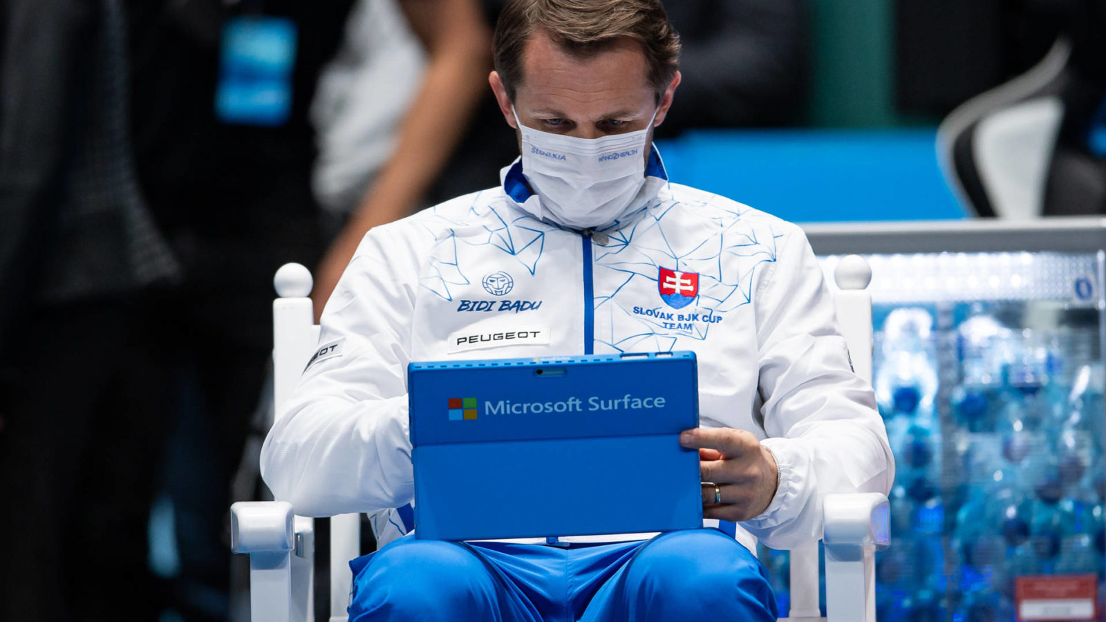 A man sits in a chair with athletic clothes and a mask on. He is holding a Microsoft Surface computer in his lap and looking at it. Various sports logos are visible on his jacket.