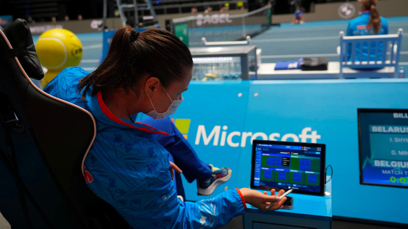 A person sits in a chair overlooking a tennis court. She is holding a Surface pen and marking it on a Surface computer. Tennis statistics appear on the screen.