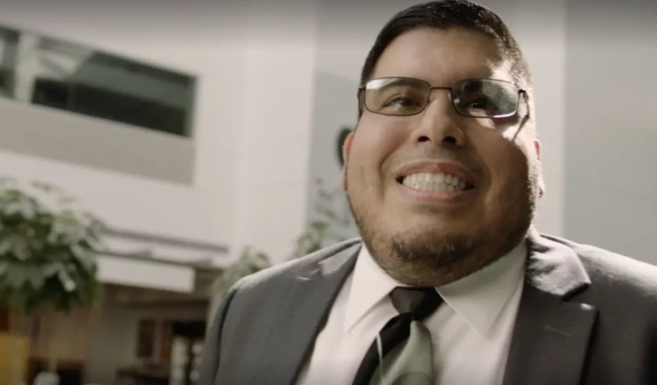A man in a shirt, tie and glasses smiling broadly in the foreground of the image. The background is slightly tilted behind him where a plant and windows are visible.