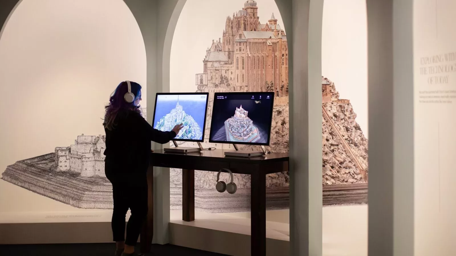 A woman dressed in black wears headphones and uses a touchscreen to explore an exhibit on the 3D model of Mont Saint Michel, on display behind arched windows in a room in front of her.