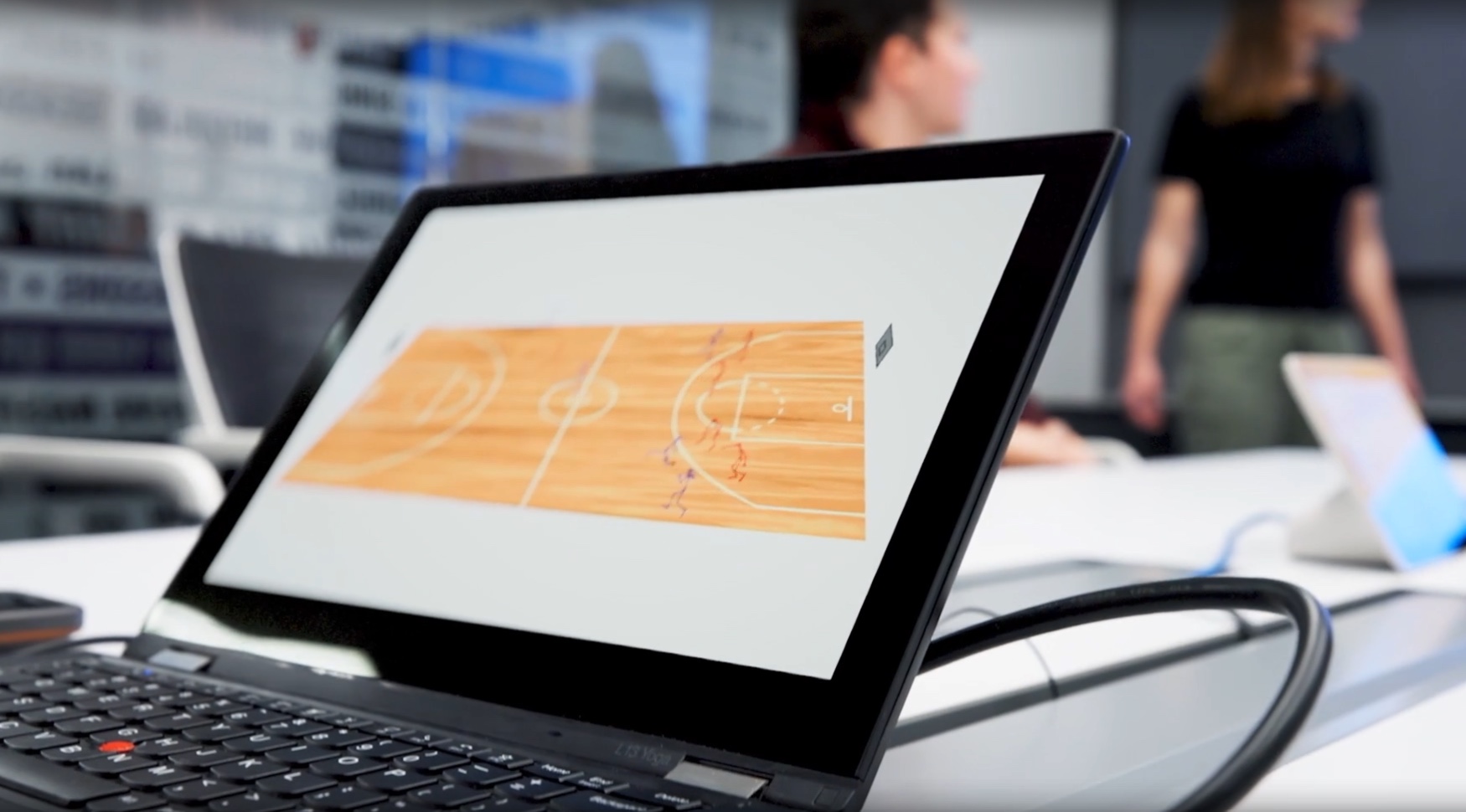 Laptop displaying a basketball court diagram with people in a meeting room in the background.