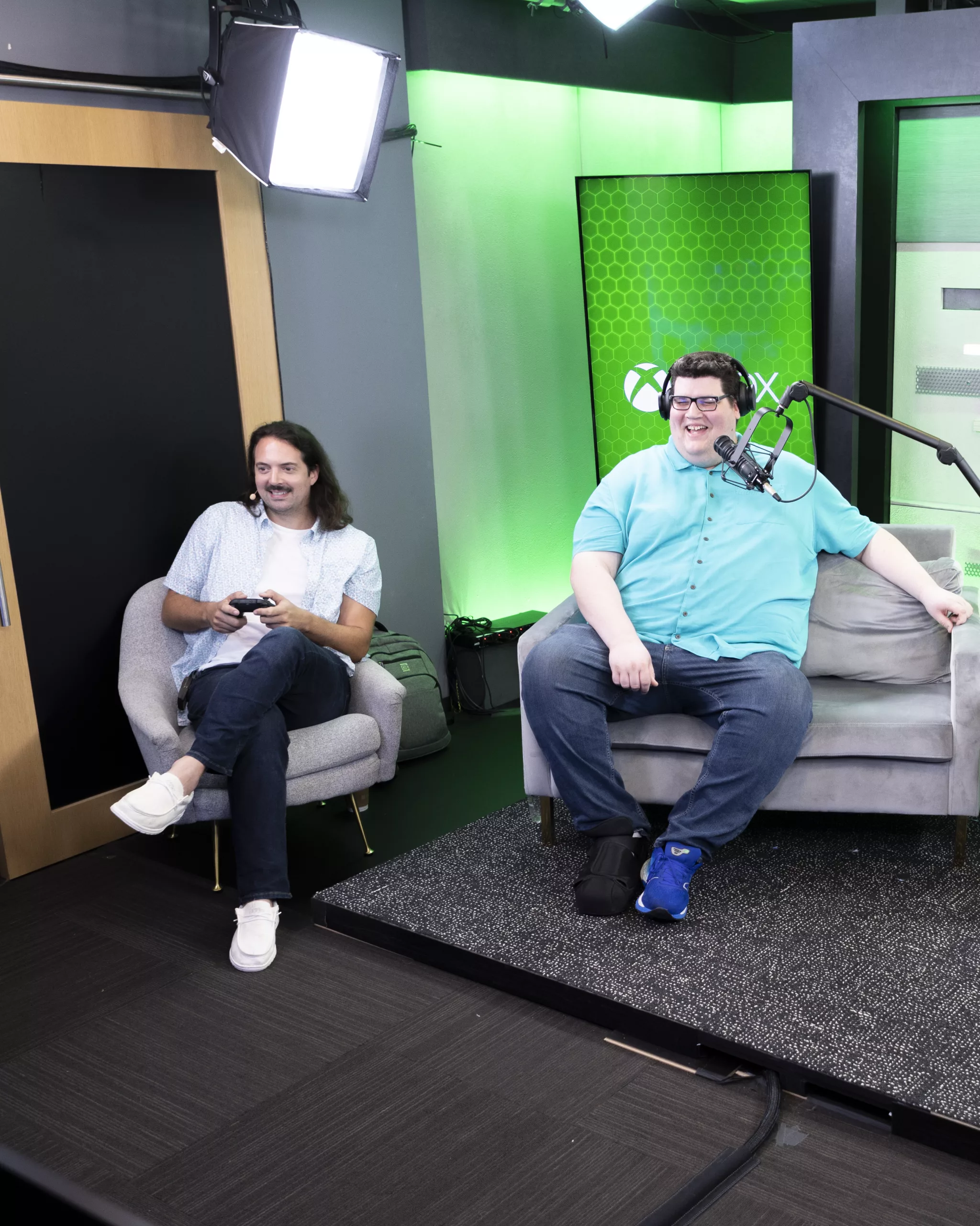 Two people sitting in chairs. One man on the left wearing a light blue shirt holds a video game controller. Another man on the left wearing a blue shirt has a microphone in front of him and is wearing headphones.