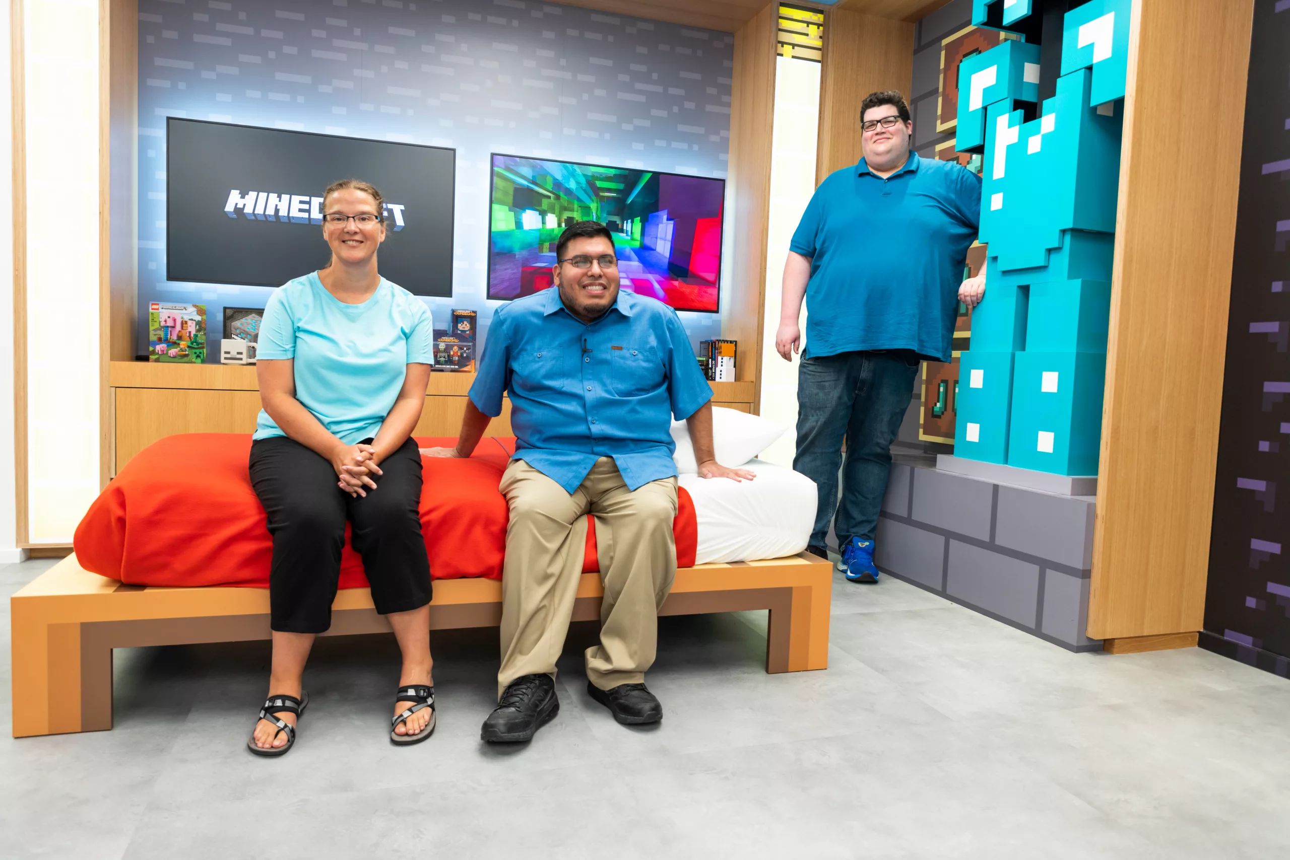 Foreground: a woman and a man sit smiling on a bed in an experiential Minecraft gaming environment while another man stands on the right next to them. Background: two screens behind the sitting people with one that says “Minecraft” and the walls are made to look like the Minecraft “building-block” aesthetic.