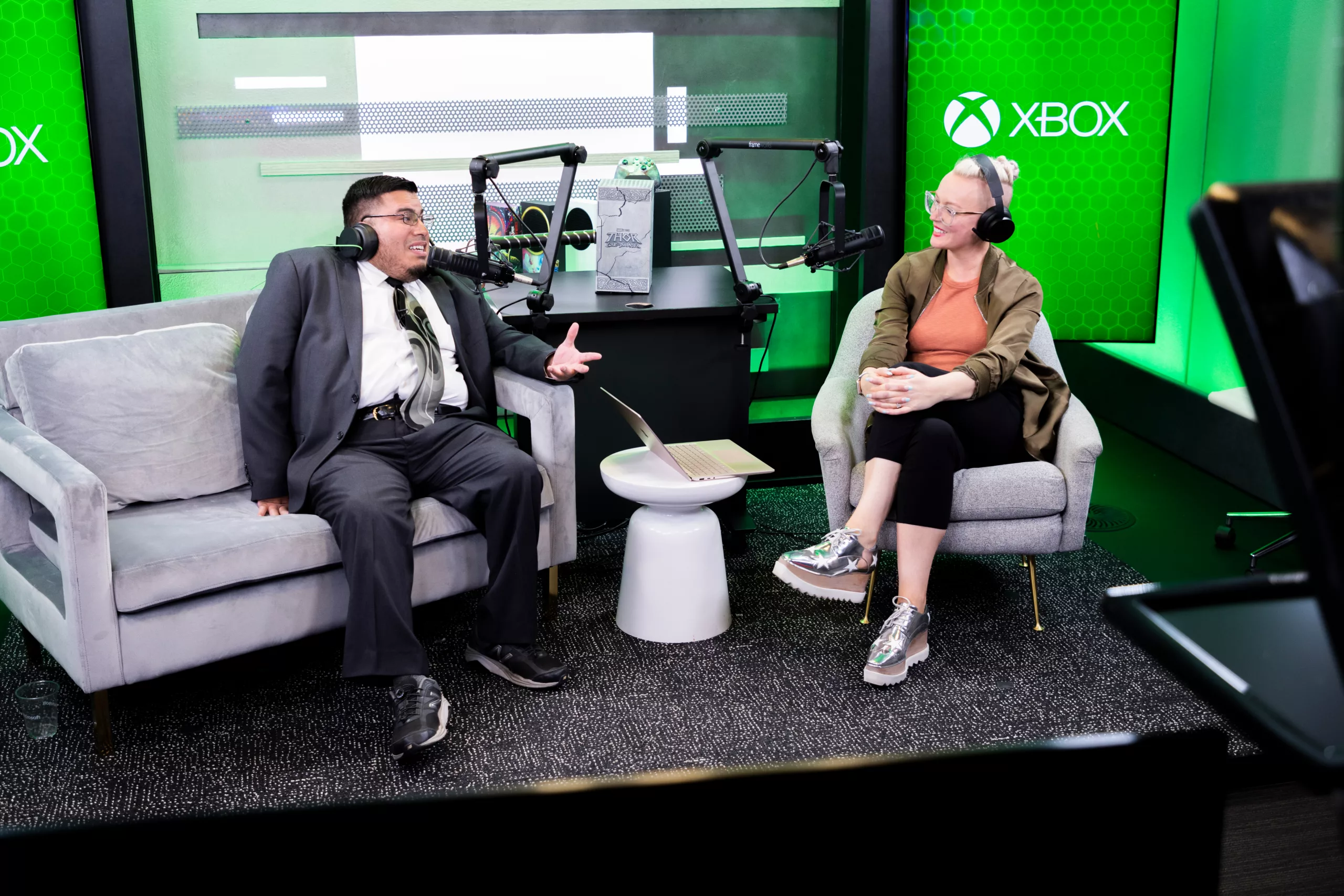 Two people sit wearing headphones. A man in a suit and tie sits on a couch on the left and a woman in an orange and brown top sits on the right. They have microphones in front of them. There are XBOX signs with XBOX logos behind them on a green wall.