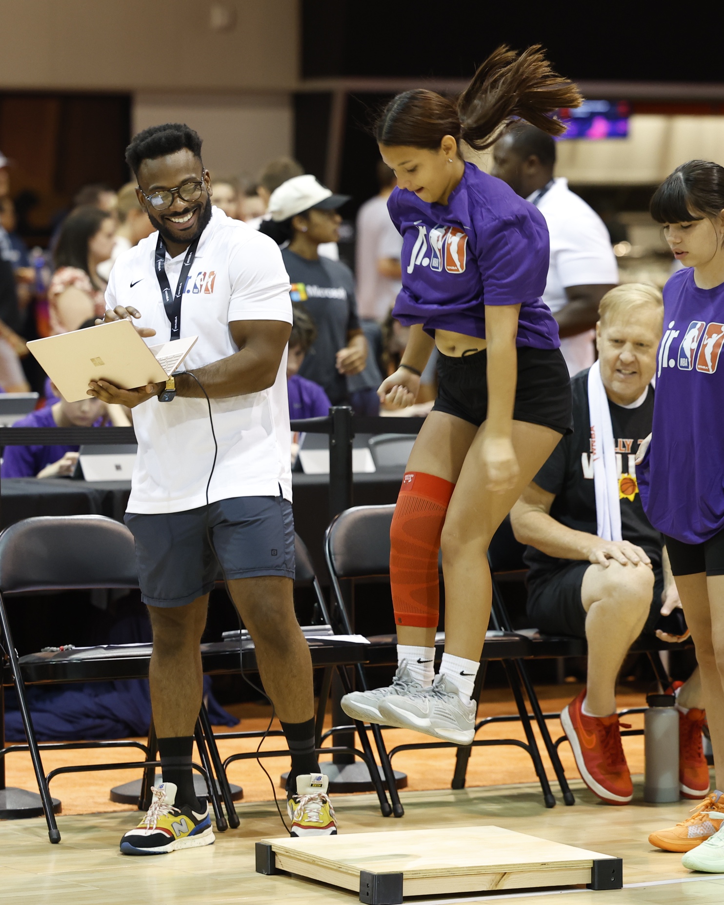 A coach records results as a young athlete in sportswear performs a vertical jump onto a platform during an indoor sports event.