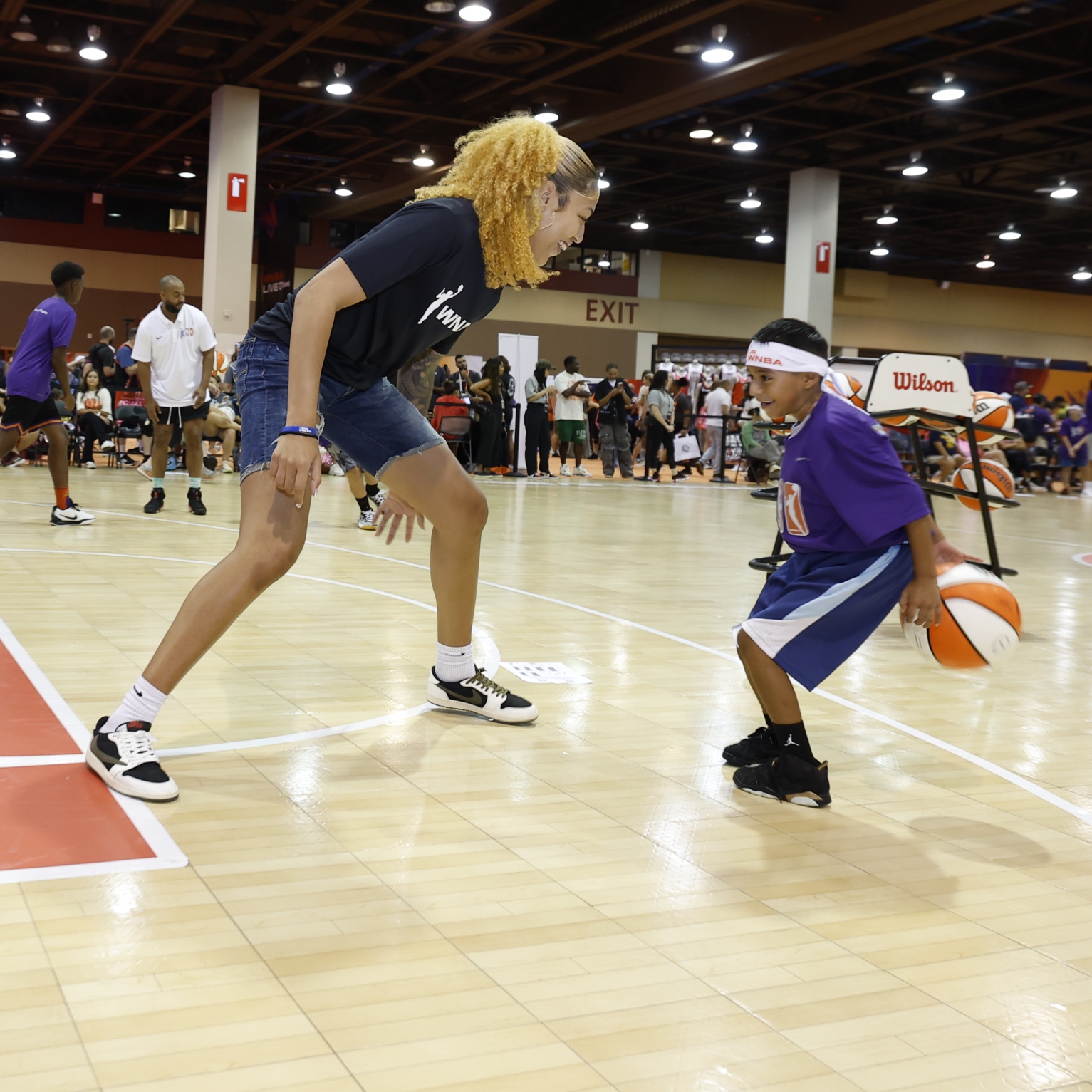 An adult and a child play basketball indoors; the adult is guarding while the child dribbles the ball, with spectators watching in the background.