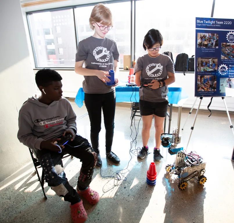 Three children look at a robot as they hold what appear to be controllers.