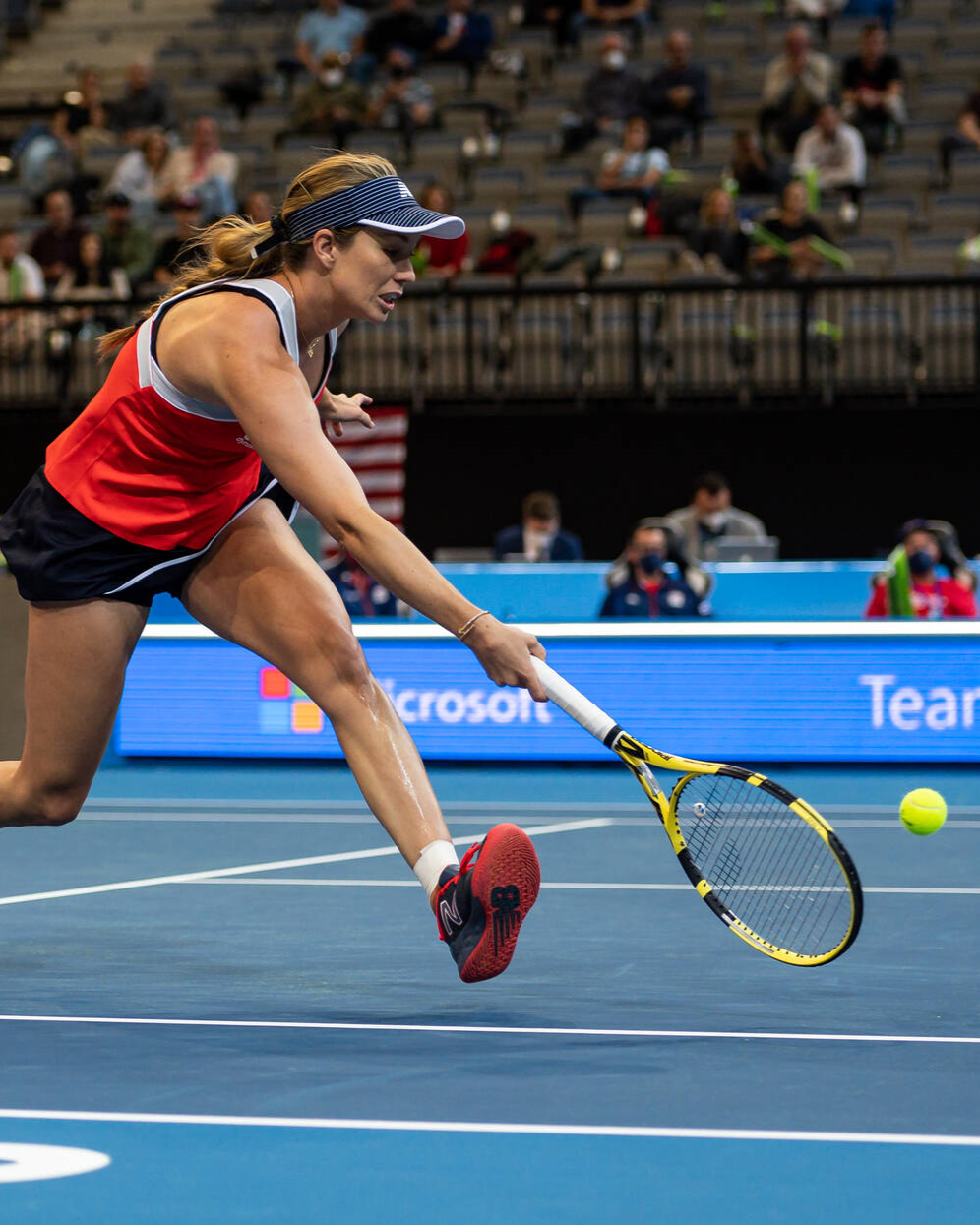 A female tennis player reaches mid-air to hit a tennis ball with her racket on the court. There are people sitting in a crowd on the stands behind her.