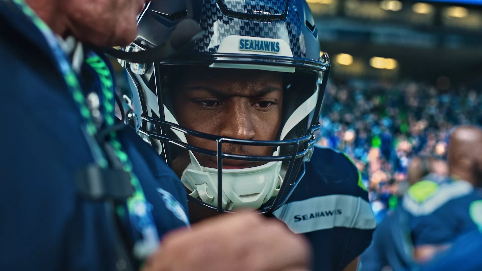 Close up portrait of NFL player Tyler Lockett looking pensive while wearing his helmet and jersey. A blurry crowd of fans are in the background.