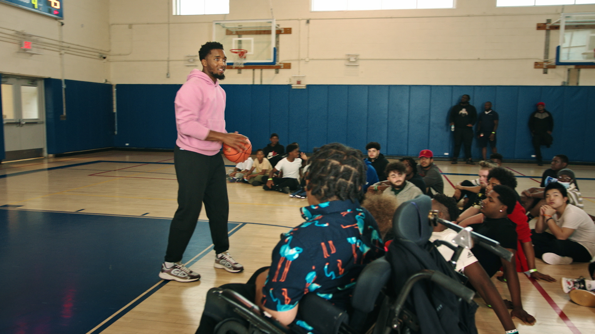 NBA star Donovan Mitchell stands in the middle of a basketball court holding a basketball. Students are seated in a semi-circle around him.