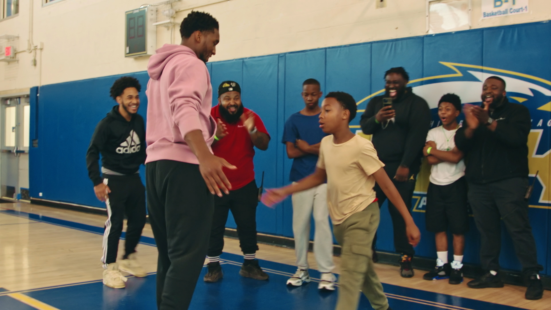 On a basketball court, an NBA basketball player is on left high-fiving a young student on the right. Four onlookers in the background smile and take pictures from their phones.