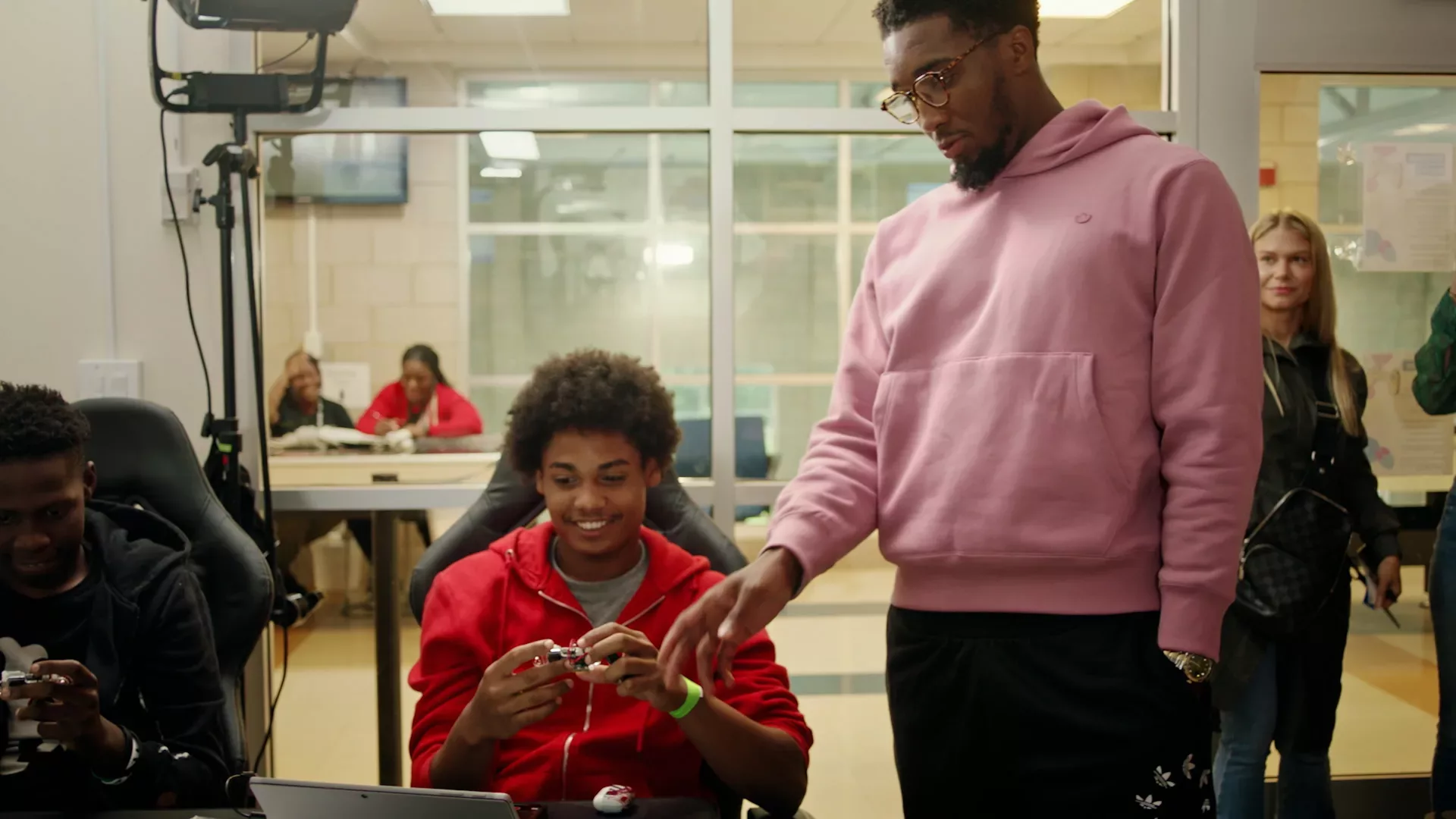 NBA star Donovan Mitchell stands over a student seated at a computer table. The student holds a computing device and is smiling.