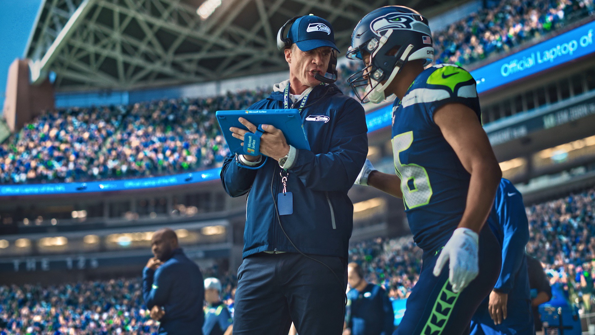 NFL player Tyler Lockett stands wearing his helmet and jersey talking to his coach who is holding a Microsoft Surface. They stand in the center frame with a crowd of fans in the stadium seats behind them.