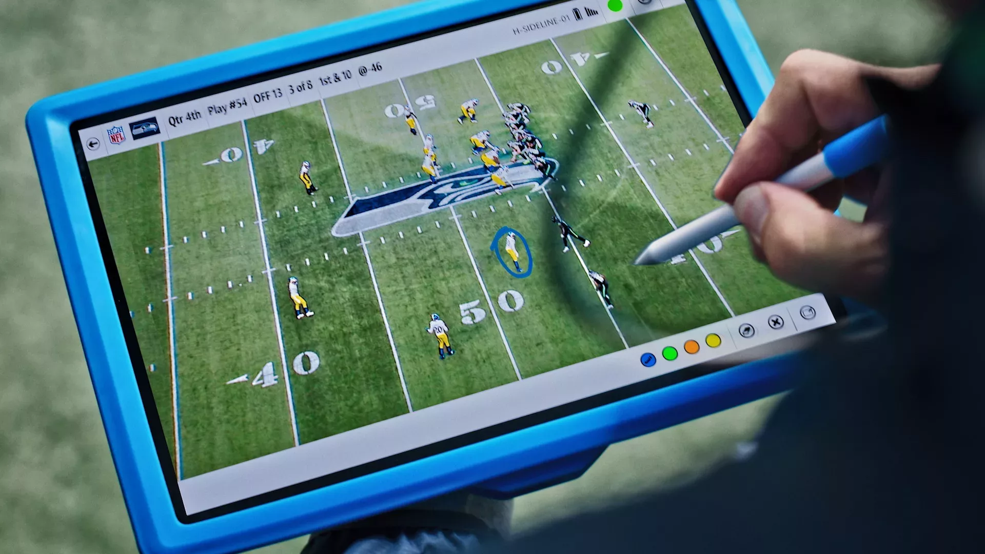 A close-up image of a Microsoft Surface with a football play visible on screen. A hand is visible in the lower right corner holding a digital stylus, making marks on the screen.