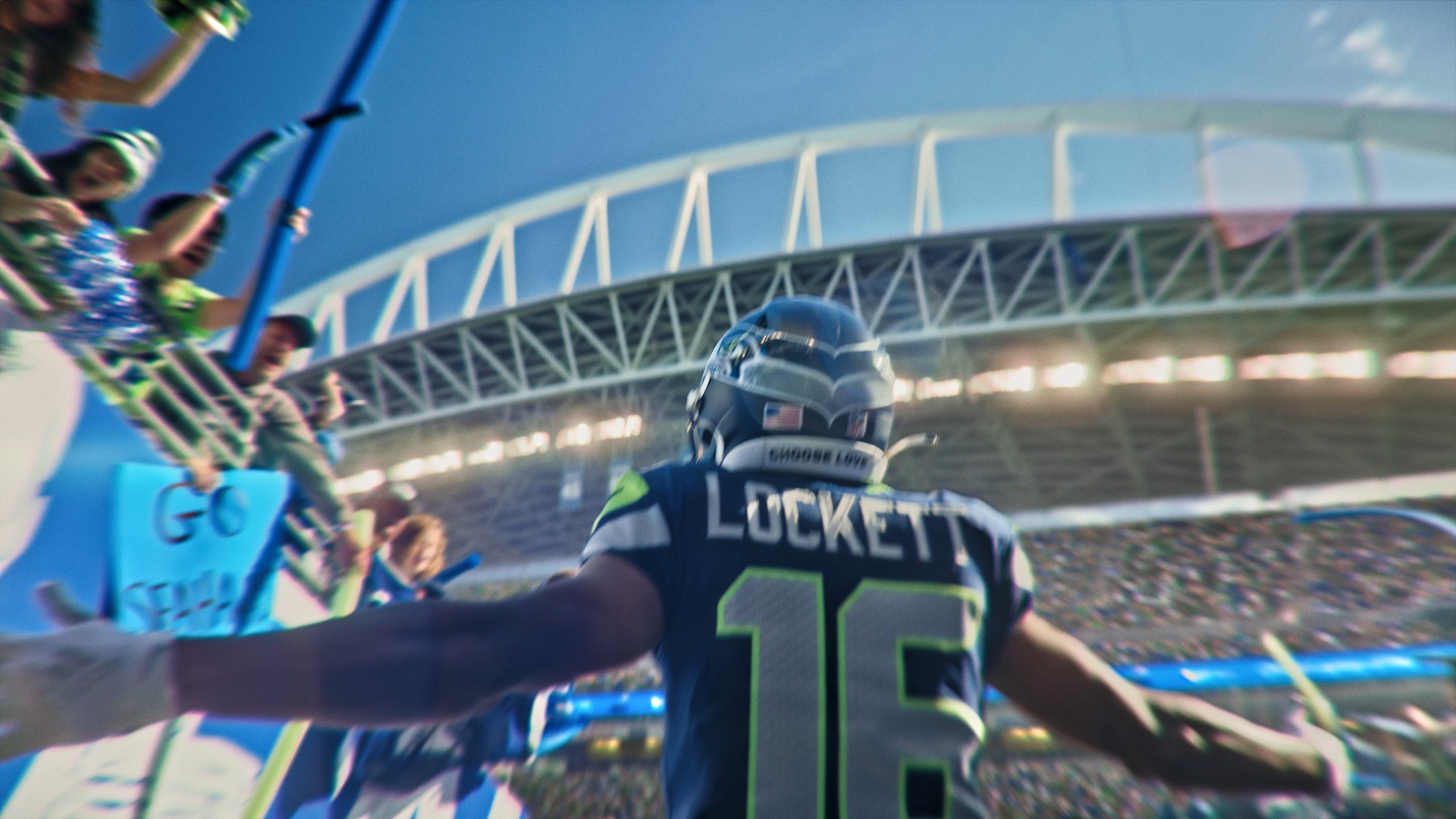 NFL player Tyler Lockett wearing a jersey and helmet shown from behind. He holds his arms open in front of a crowded stadium of fans. The back of his jersey says “LOCKETT 16.”