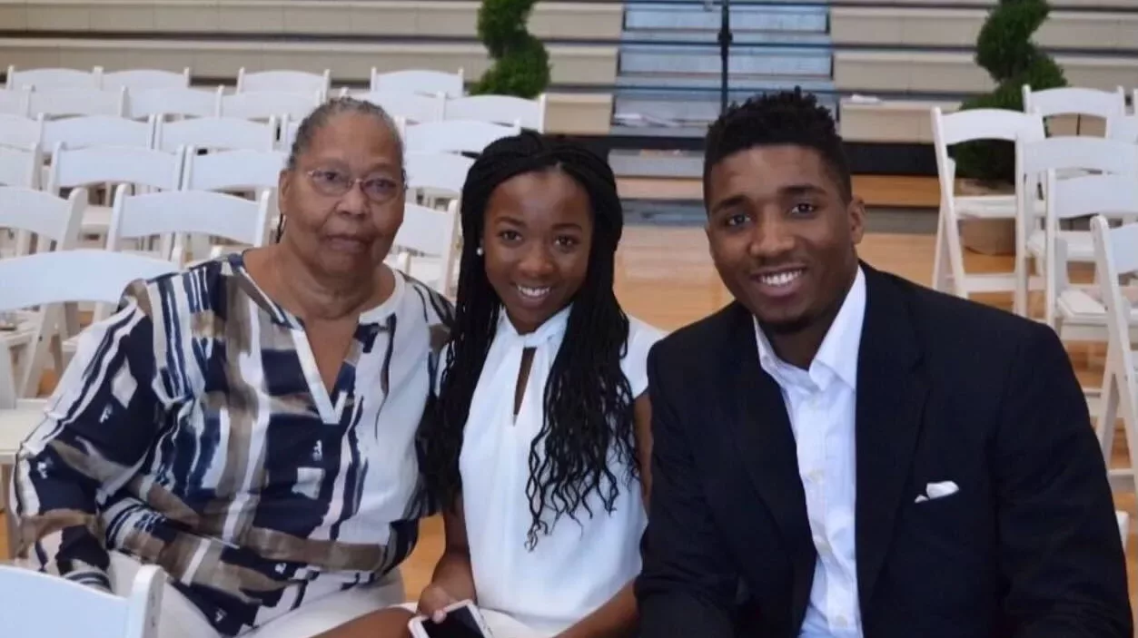 Three people sit smiling in the middle of a basketball court with empty white chairs behind them. From right to left, the people seated are NBA star Donovan Mitchell, his sister and his grandmother.