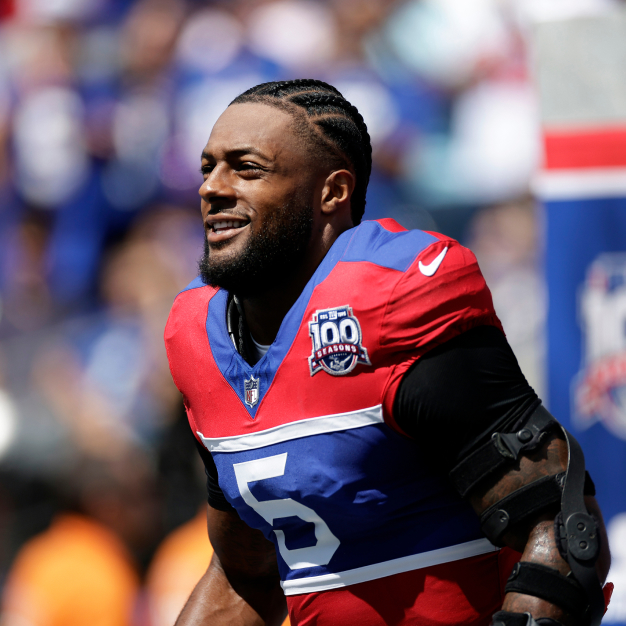 Football player in a red and blue jersey with the number 5 smiles on the field. He's wearing a large arm brace and has braided hair. Crowd in the background.