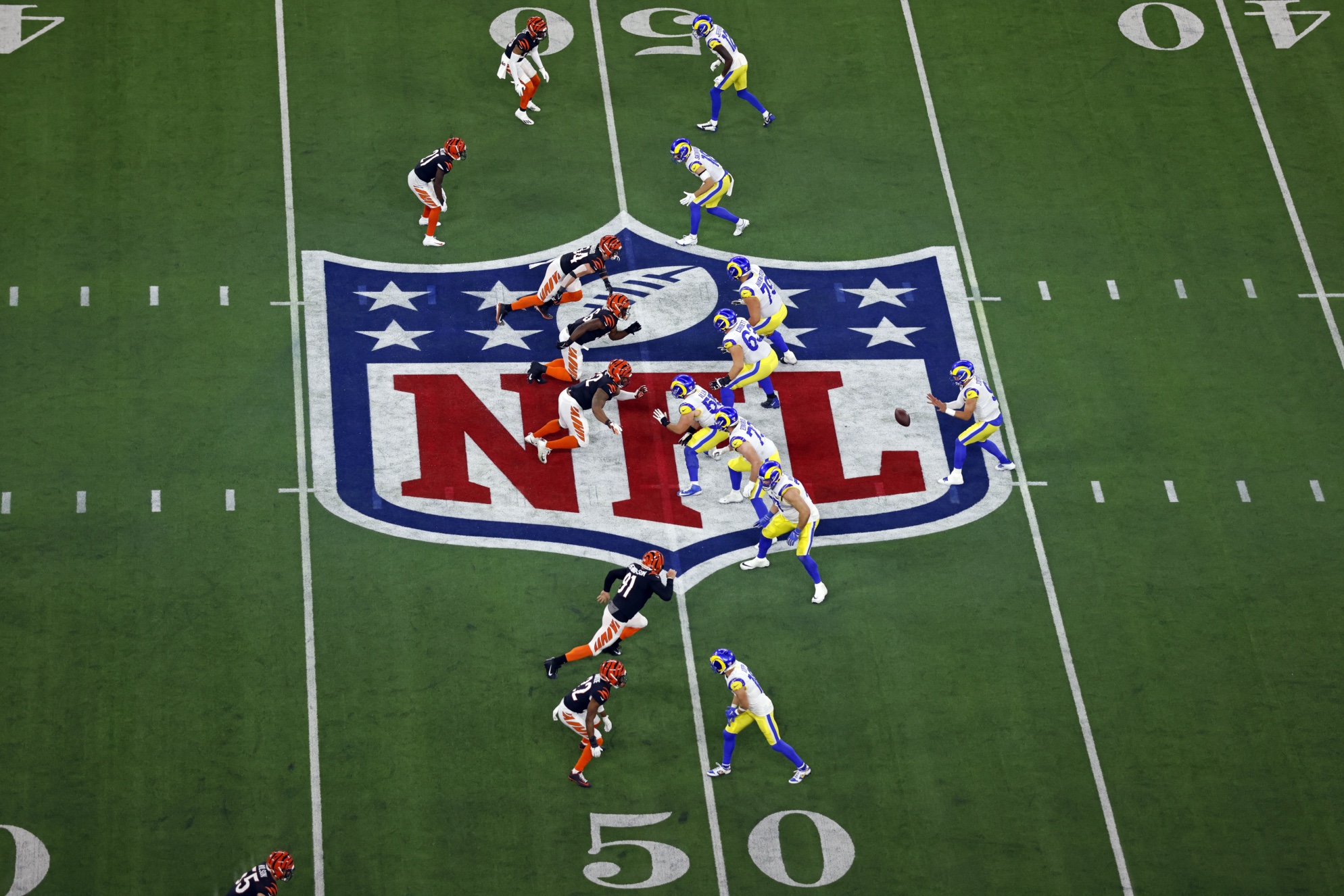 NFL players from two teams, one in orange and black and the other in yellow and blue, line up on the field near the 50-yard line, with the NFL logo visible at midfield.