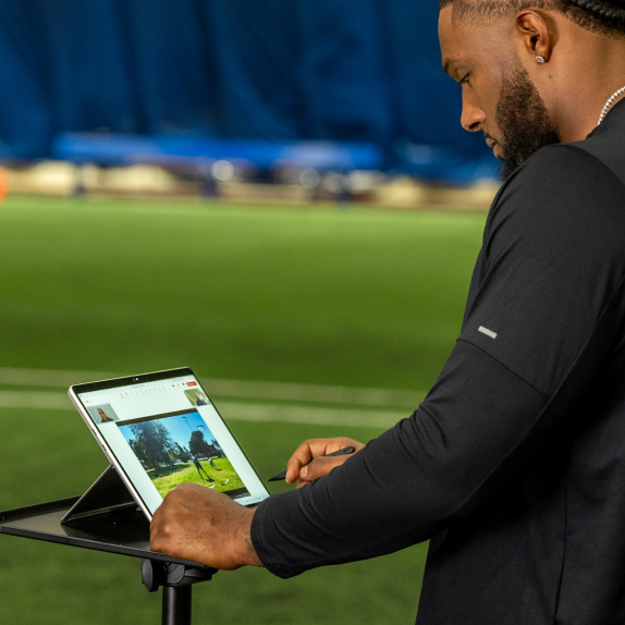 A person using a tablet on a stand, positioned on a sports field with a blue backdrop.