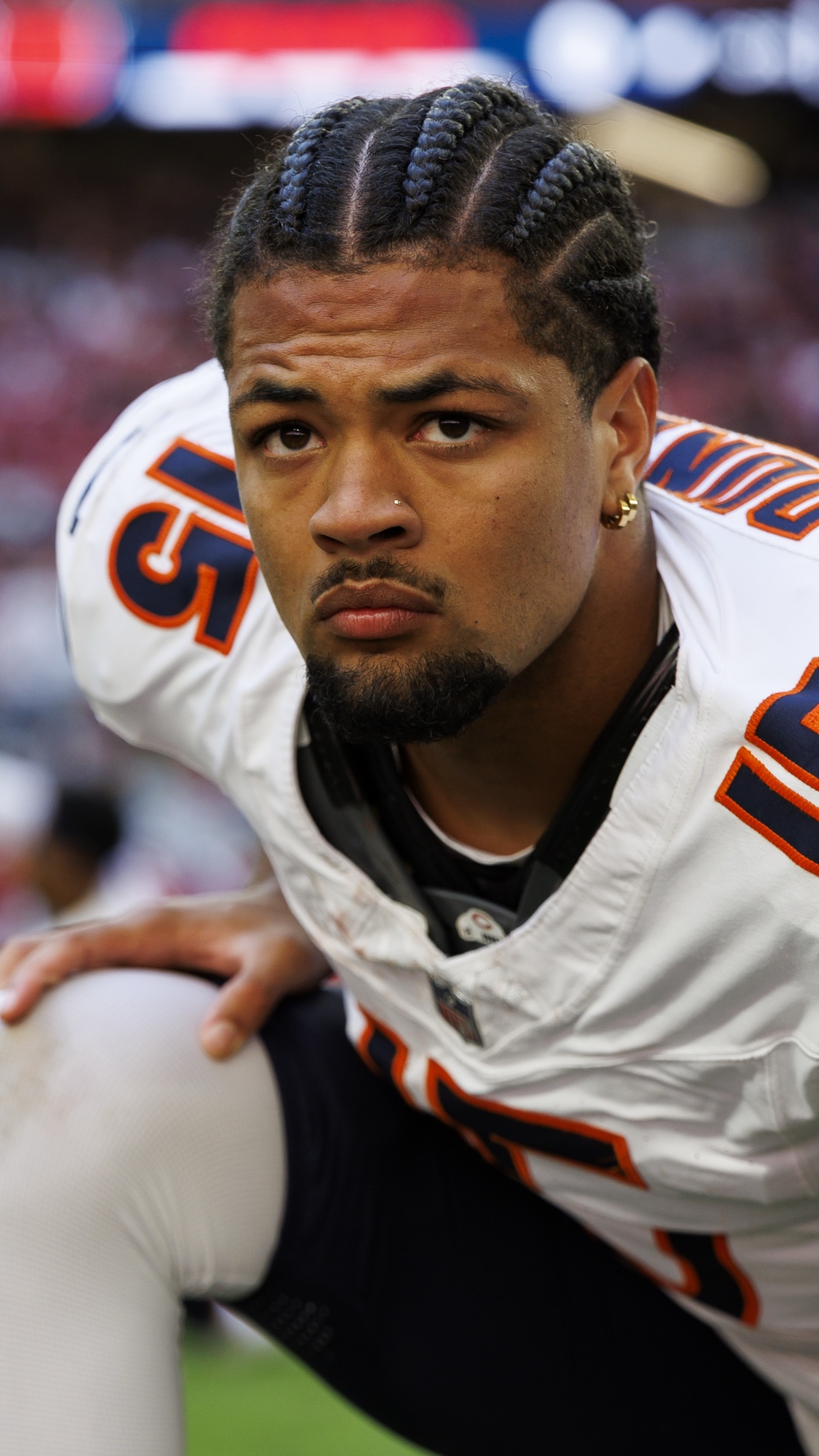 A football player in a white jersey with the number 15 and cornrow hairstyle is crouching on the field, appearing focused.