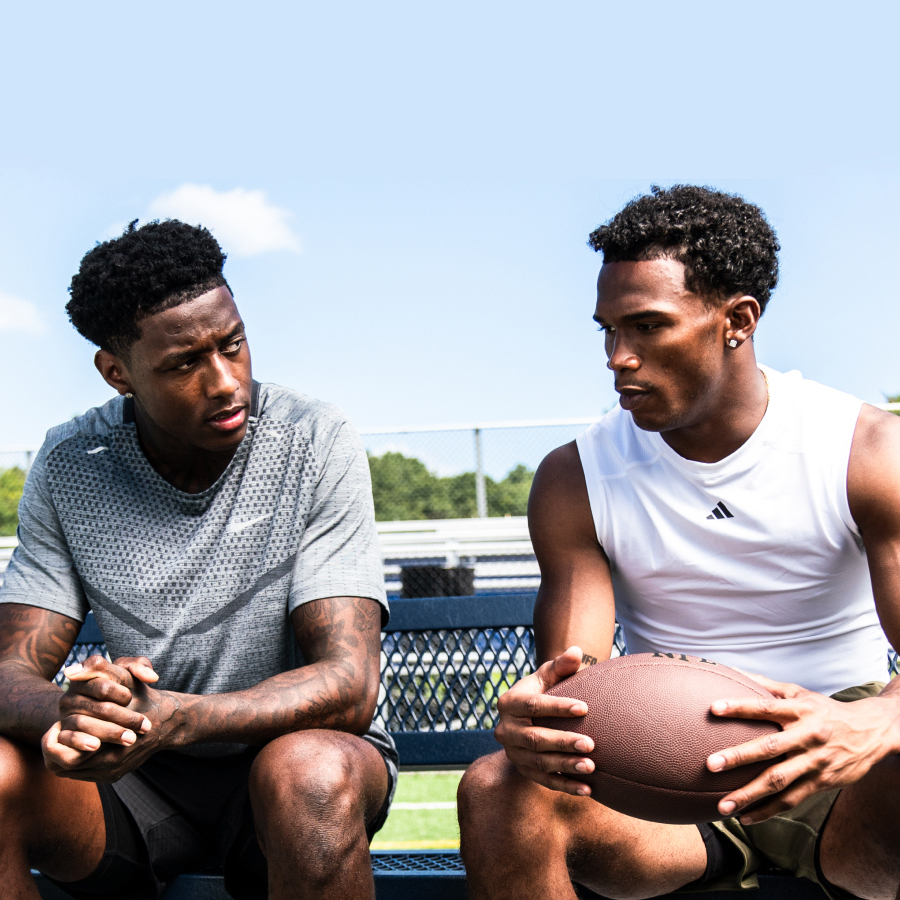 Two young men sit on metal bleachers outdoors, one in a gray shirt and the other in a white sleeveless shirt holding a football. They appear to be having a serious conversation on a sunny day.