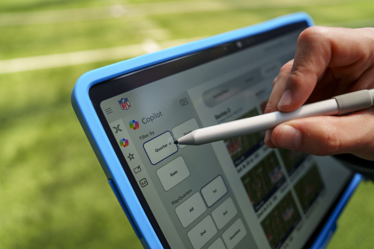 A hand uses a stylus to interact with a tablet displaying an NFL app interface, featuring a filter menu and football play images, with a grassy field blurred in the background.