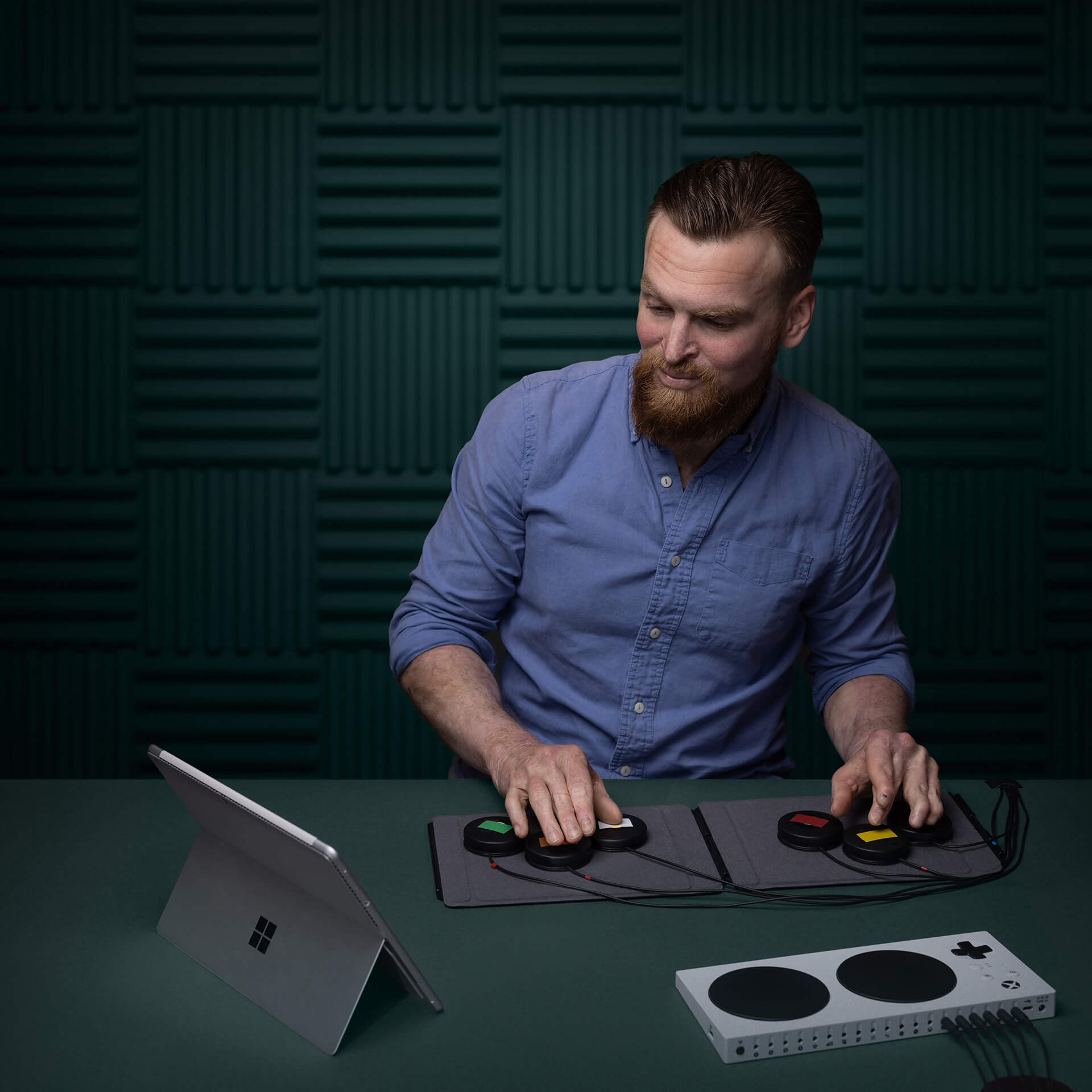 A man with a beard uses adaptive gaming equipment, including a controller with large buttons, connected to a tablet on a desk in a dark room with soundproofing panels on the walls.