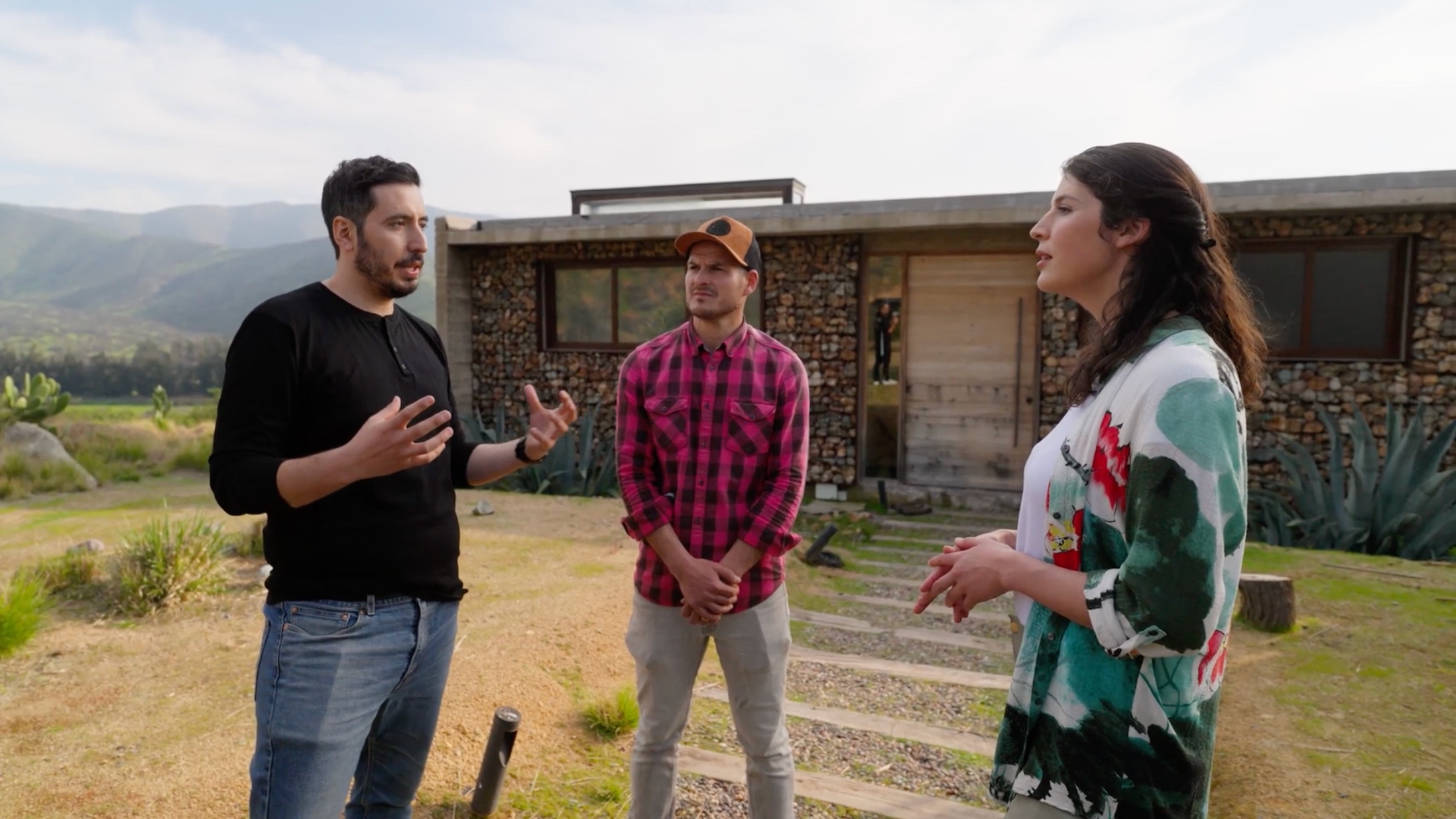 Three people stand outside a stone house, engaged in a conversation. The man on the left gestures with both hands.