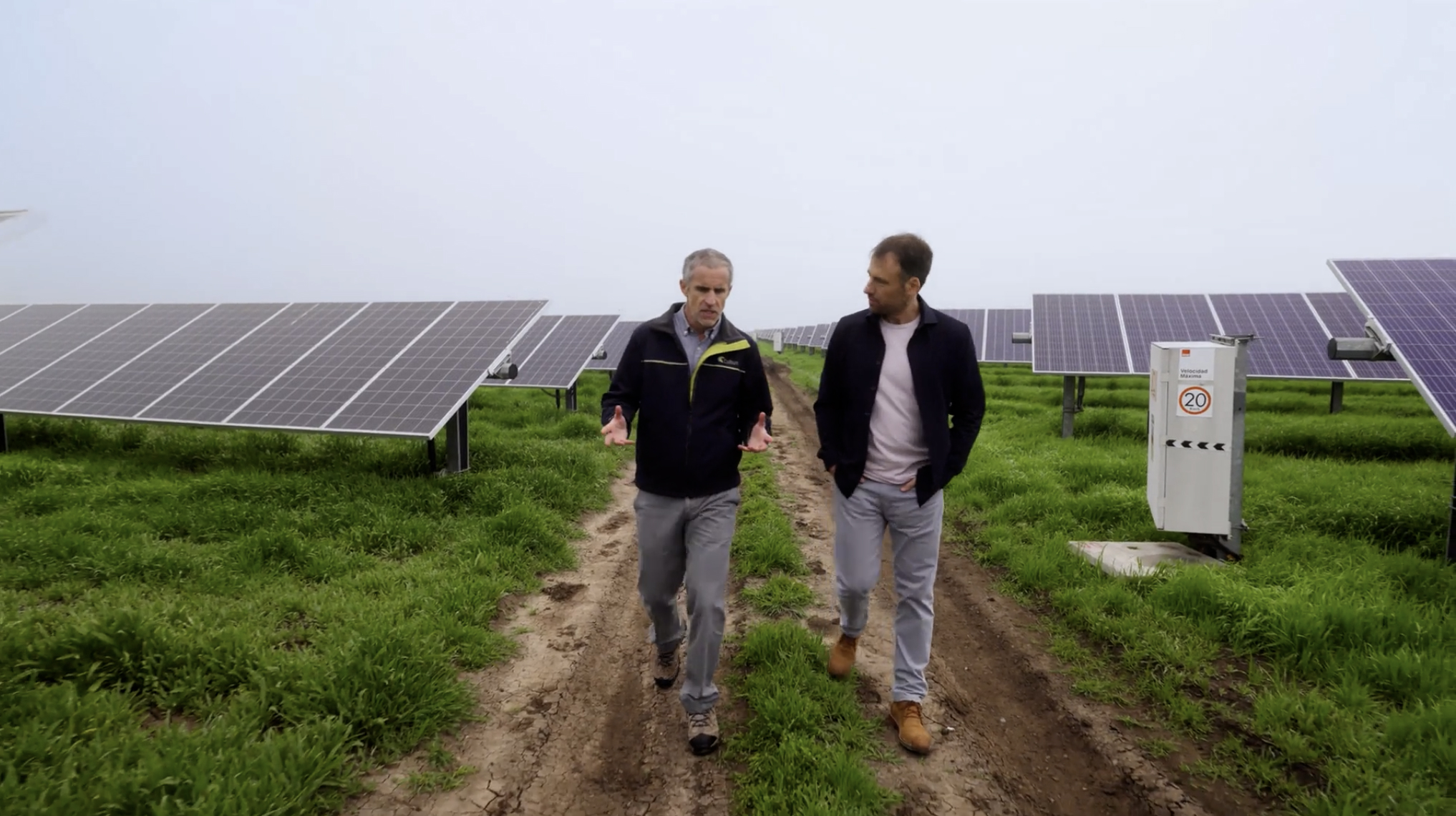 Two men walk and talk on a dirt path surrounded by solar panels in a grassy area.