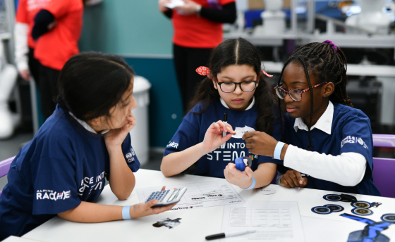 Three young girls wearing blue STEM-themed shirts work together on a project. One girl holds a calculator, and another examines a small object while referring to a paper.