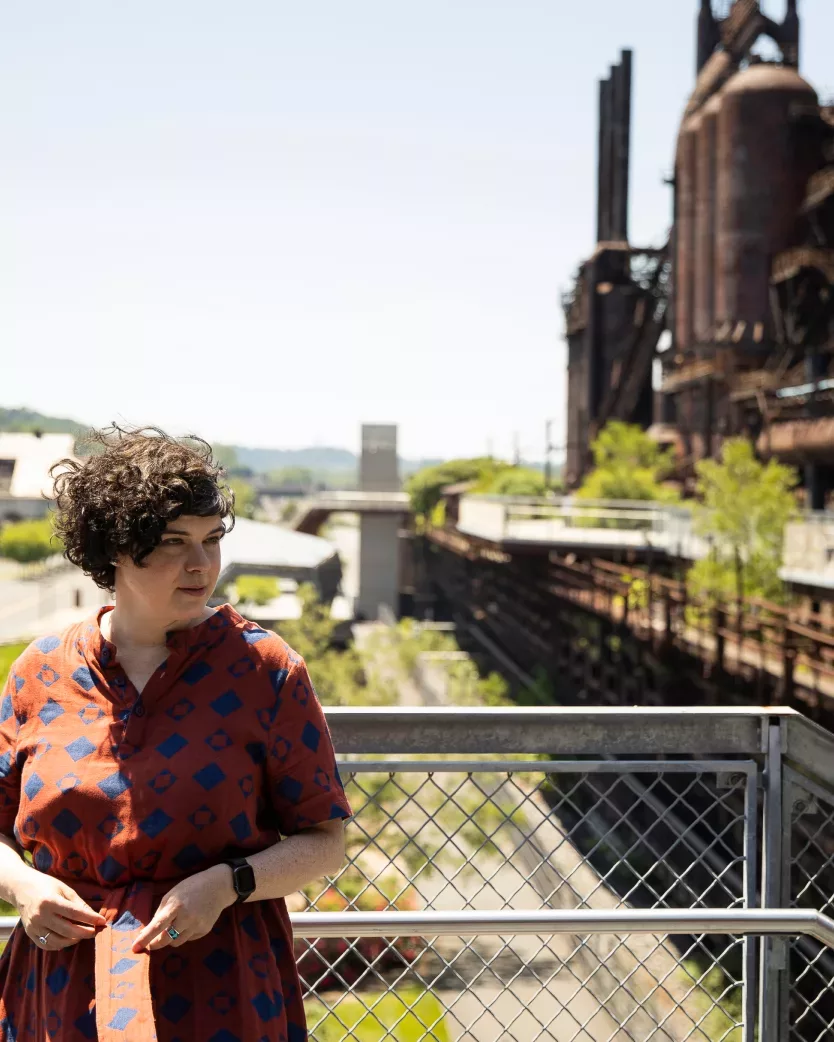 Sarah Krasley stands in front of a wire fence in Allentown, PA. Old factory buildings of Bethlehem Steel are in the background behind her.