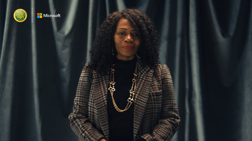 Bonita Hampton Smith wearing a black top, plaid blazer, and a gold necklace standing and looking at the camera. There is a black glossy curtain in the background.