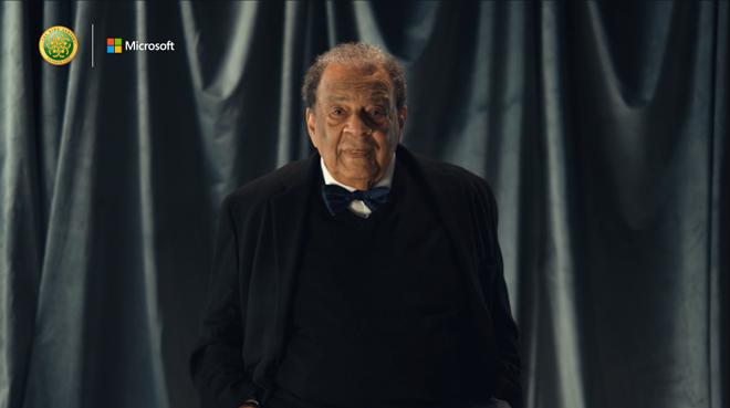 Ambassador Andrew J. Young wearing a black blazer and a bow tie while standing and looking at the camera. There is a black glossy curtain in the background.