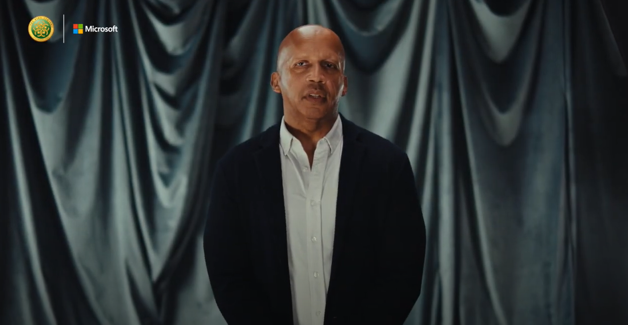 Bryan Stevenson wearing a button-down shirt and a black coat while standing and looking at the camera. There is a black glossy curtain in the background.