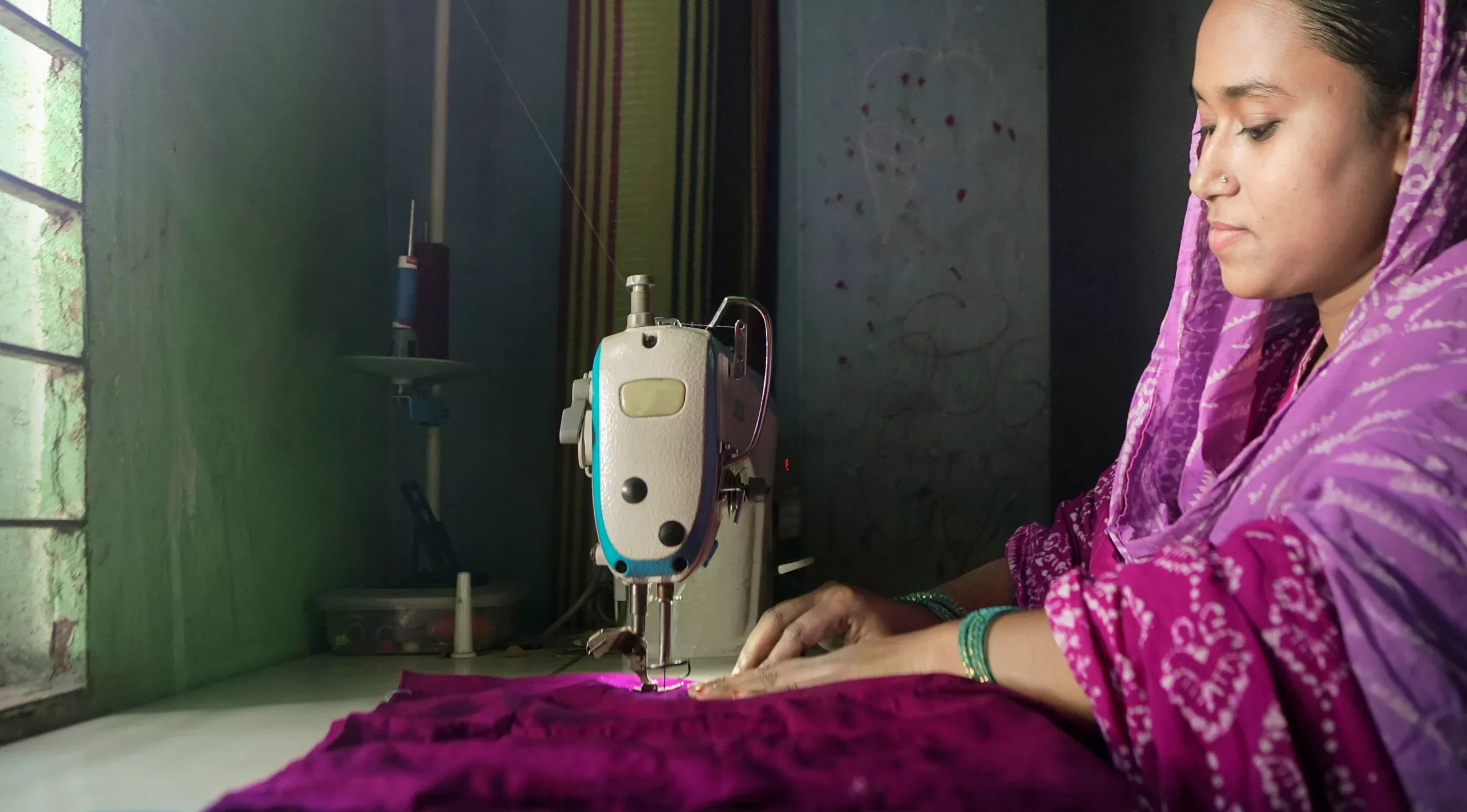 Ruma Akter sits while using her sewing machine. She is inside her home in front of a large window.
