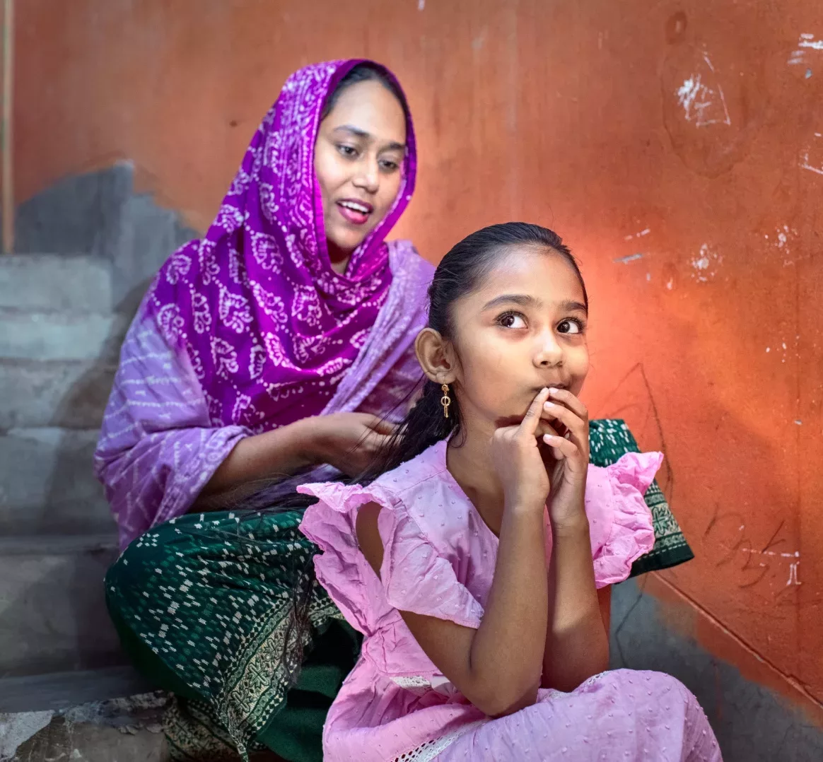 Ruma Akter braids her daughter’s hair while they both sit outside on steps.