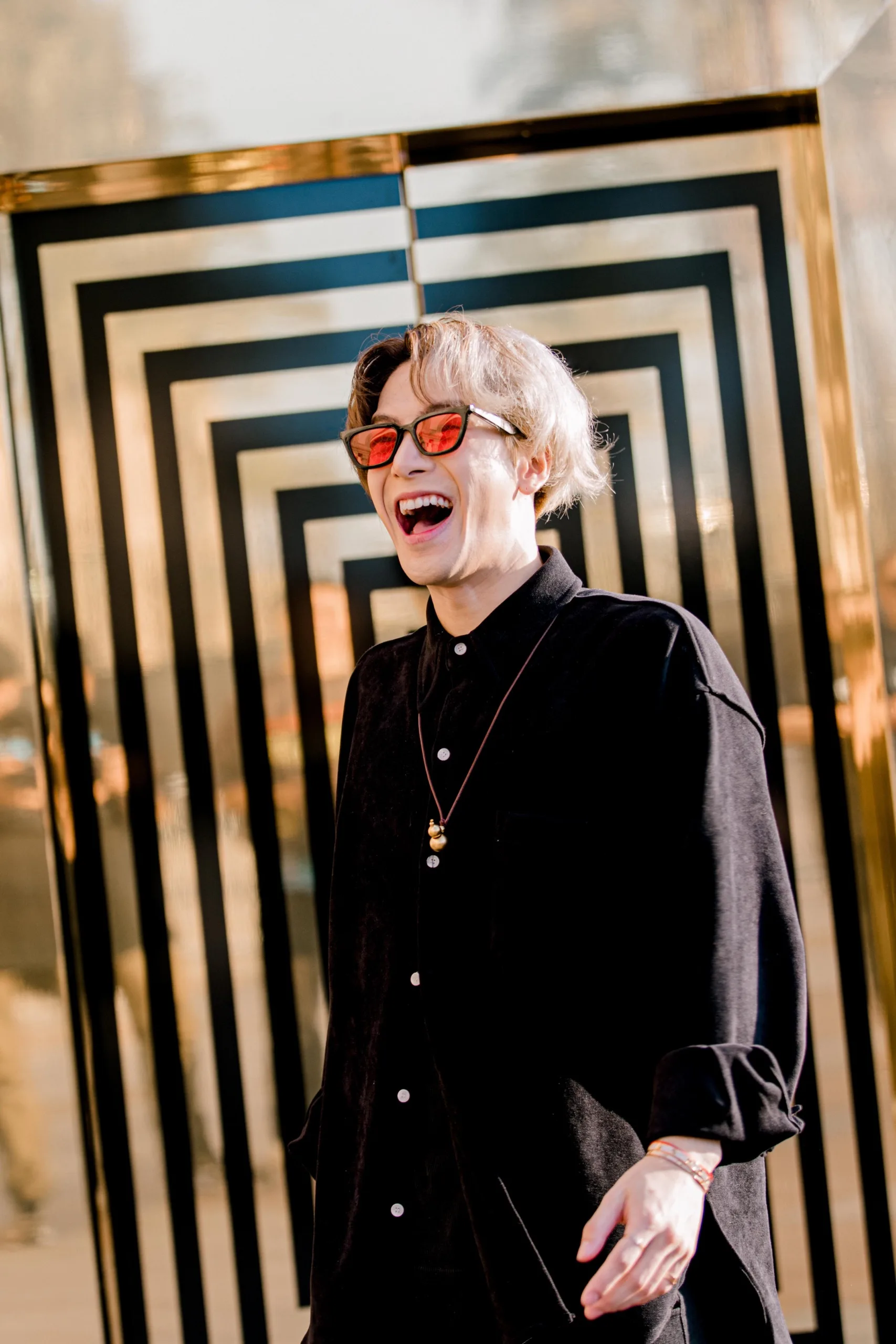 Jackson Wang pictured in a black shirt and glasses with colored lenses as he smiles in front of a shiny gold and black patterned background.