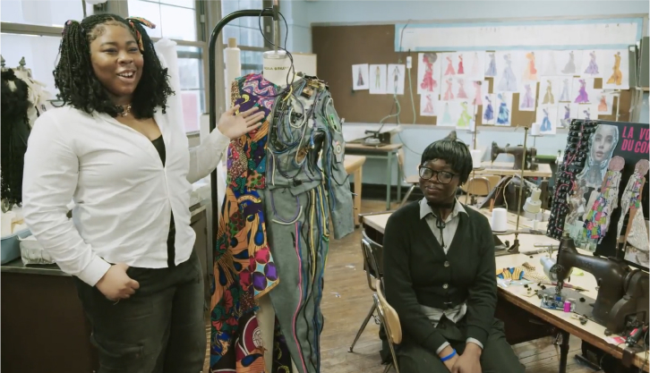 Two individuals in a fashion design studio, one standing by a mannequin draped with vibrant fabric, the other seated near a sewing machine.